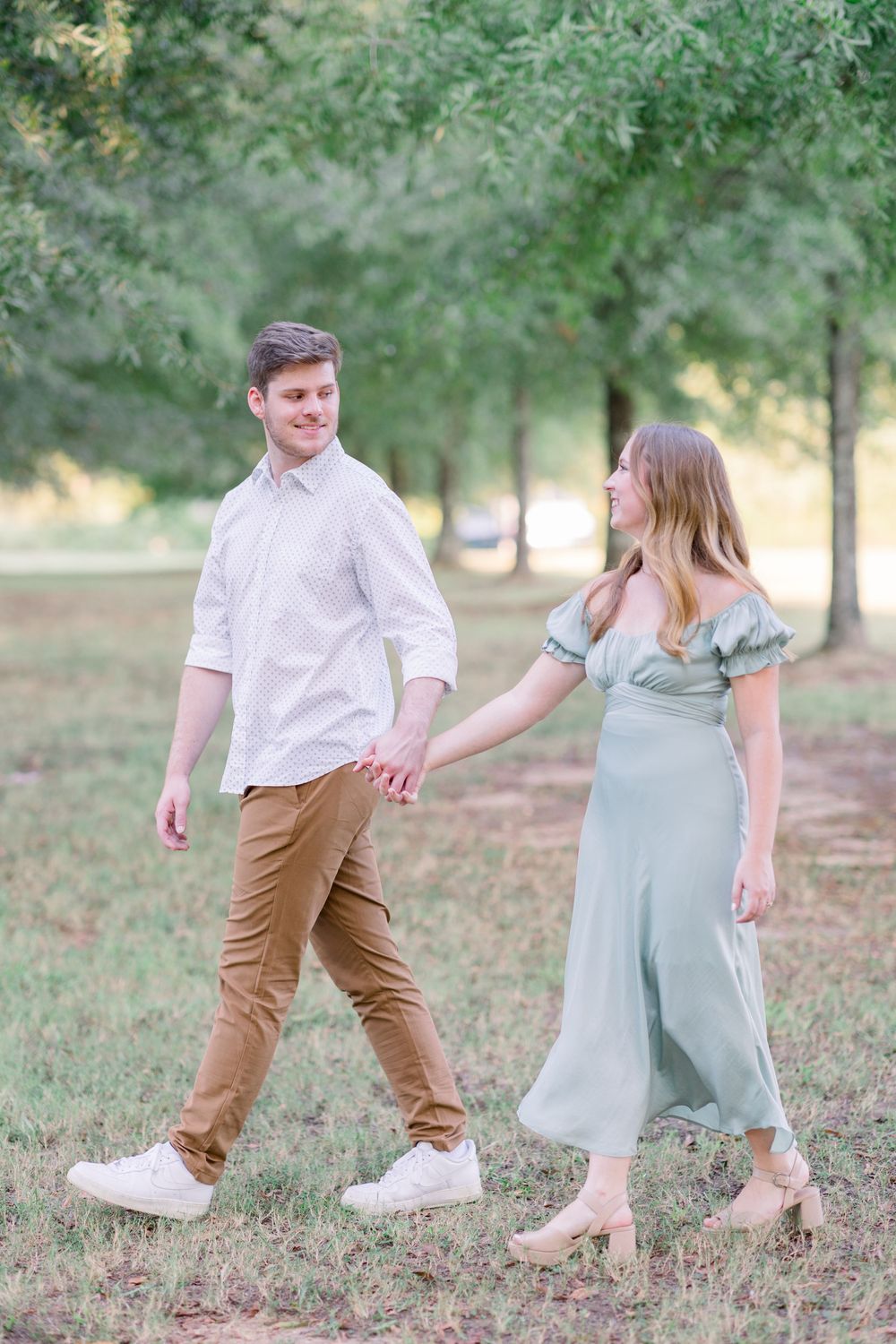 A couple walks hand in hand through a wooded park, wearing casual attire on a sunny afternoon.