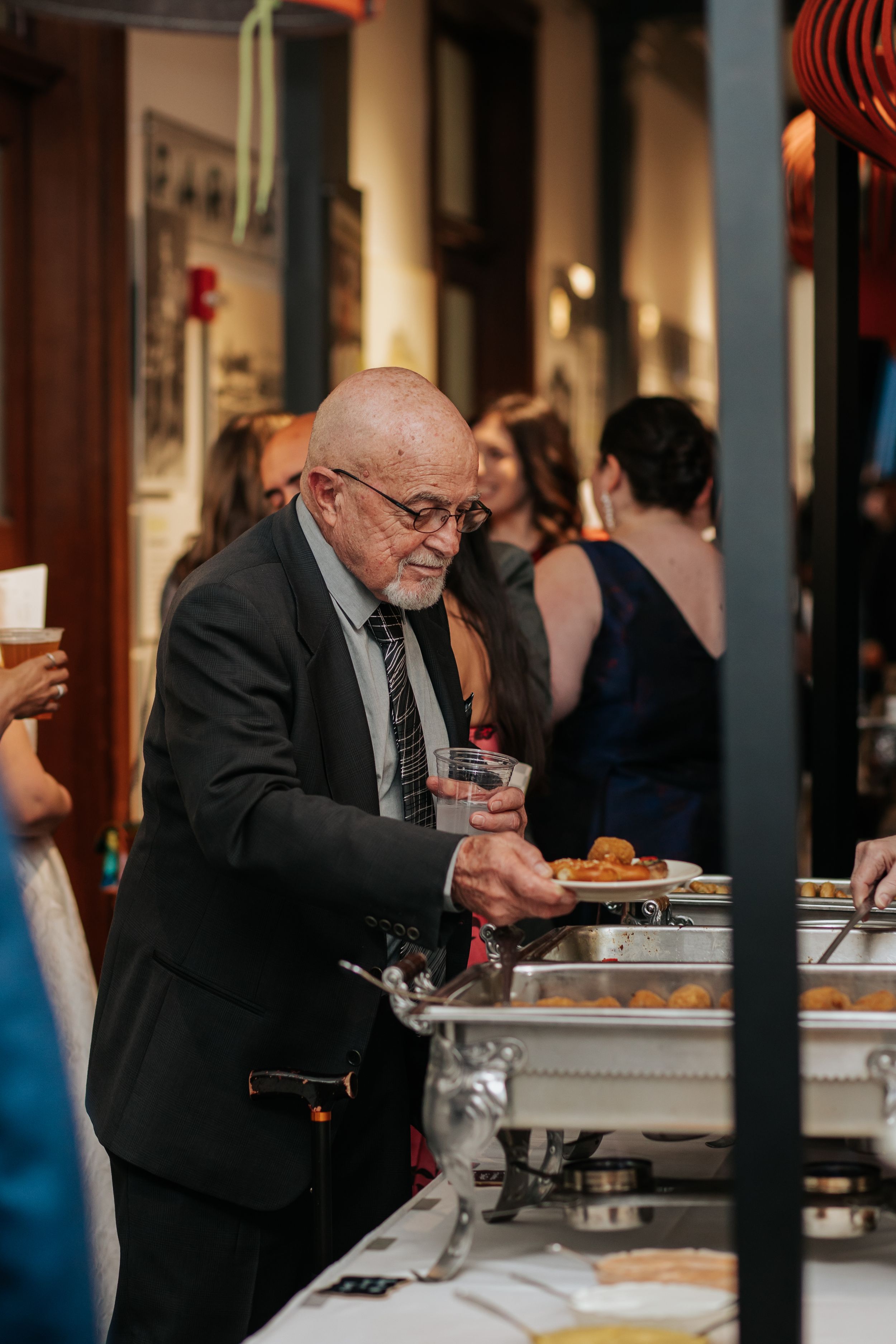 An elderly man in a dark suit serves himself food from a buffet line at an indoor event.
