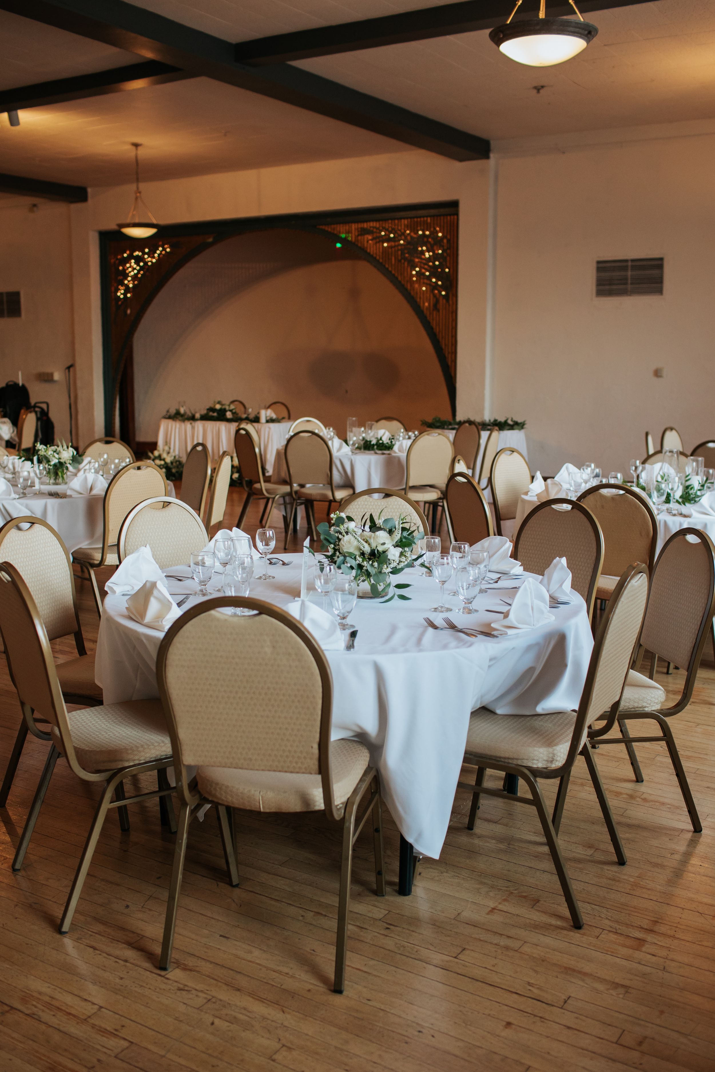 Elegant banquet hall setup with round tables covered in white linens and floral centerpieces on hardwood floors.