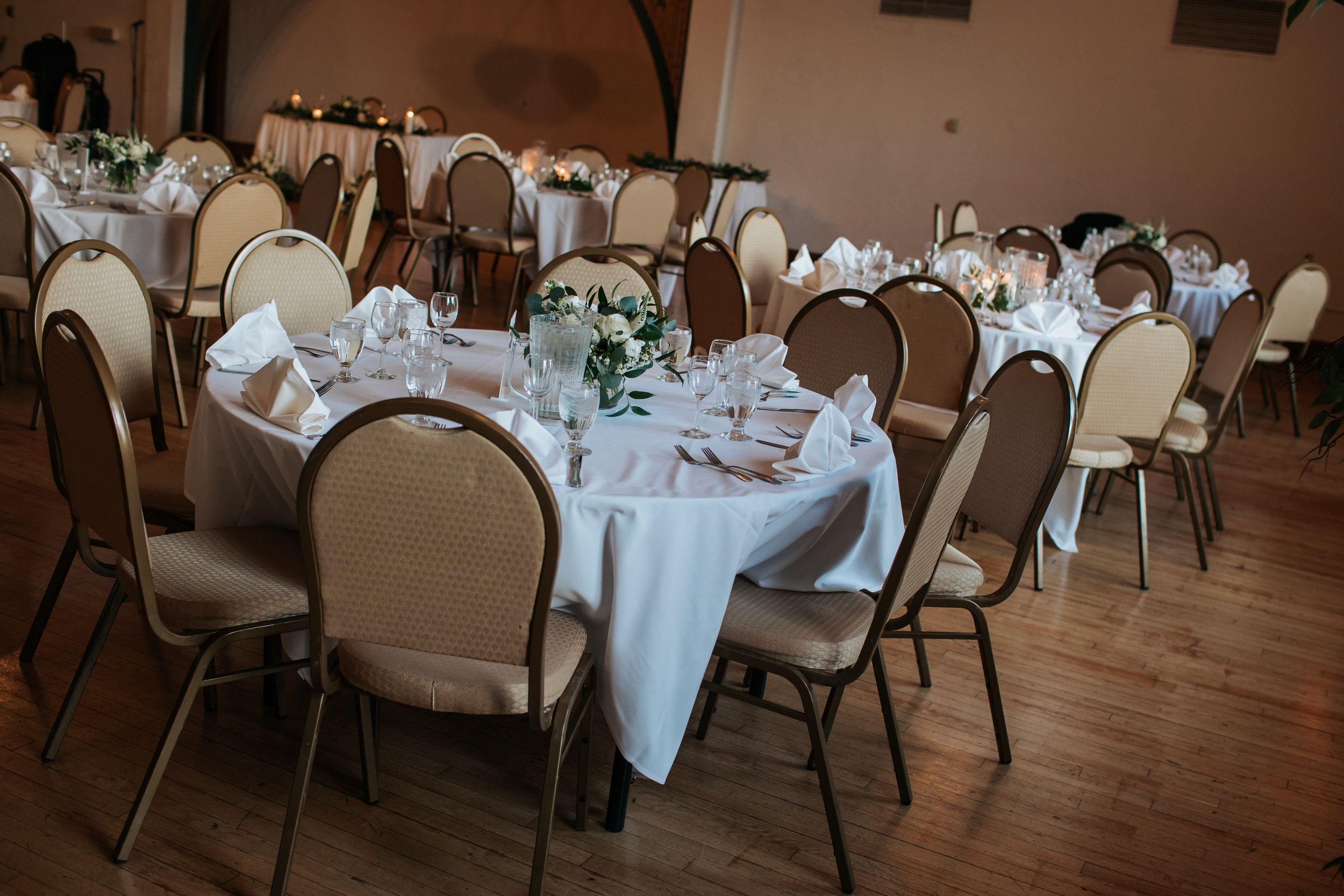 Elegant wedding reception venue with round tables draped in white linens and padded chairs arranged on wooden flooring.