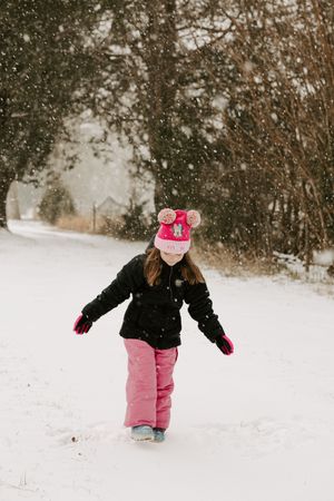 Small child in pink snow pants and black jacket playing in snowy winter wonderland during snowfall.
