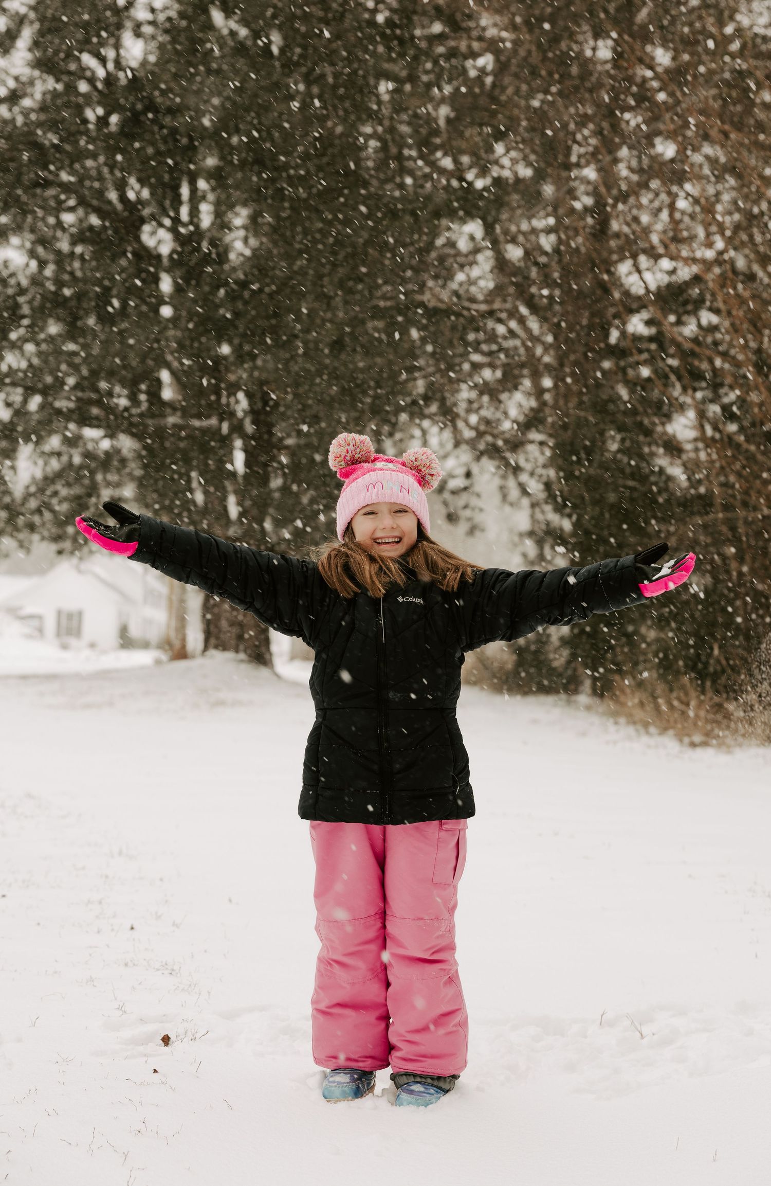 A child in a black winter coat and pink snow pants plays joyfully in the falling snow.