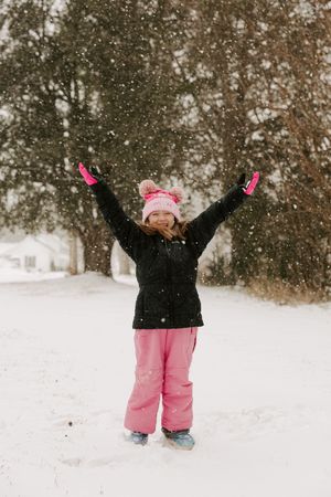 A child in pink snow pants and black jacket plays joyfully in falling snow with arms raised up to the sky.