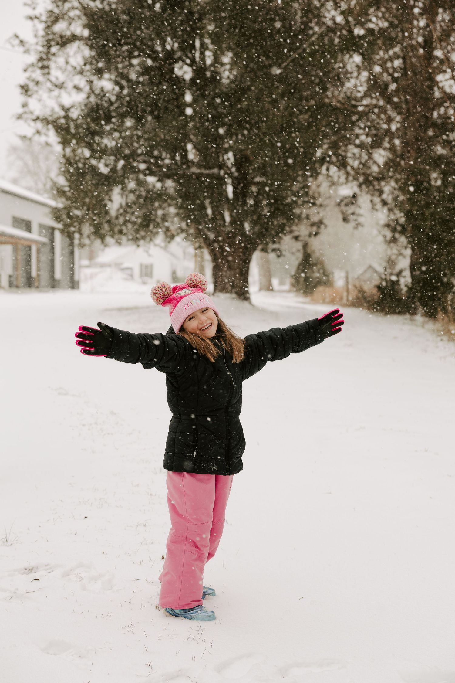 A child in pink snow pants and black jacket plays in the snow during winter, with trees in the background.