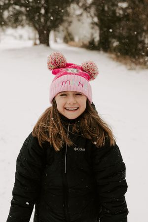 A person wearing a pink winter hat and black coat smiles while enjoying a snowy winter day outdoors.