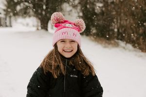 Pink winter hat with pom poms and black coat in snowy outdoor setting during snowfall.