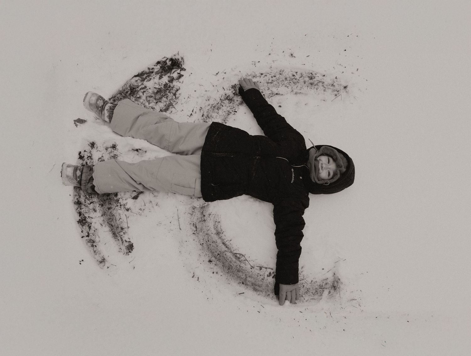 A person in black clothing lies in the snow making a snow angel against a white winter background.