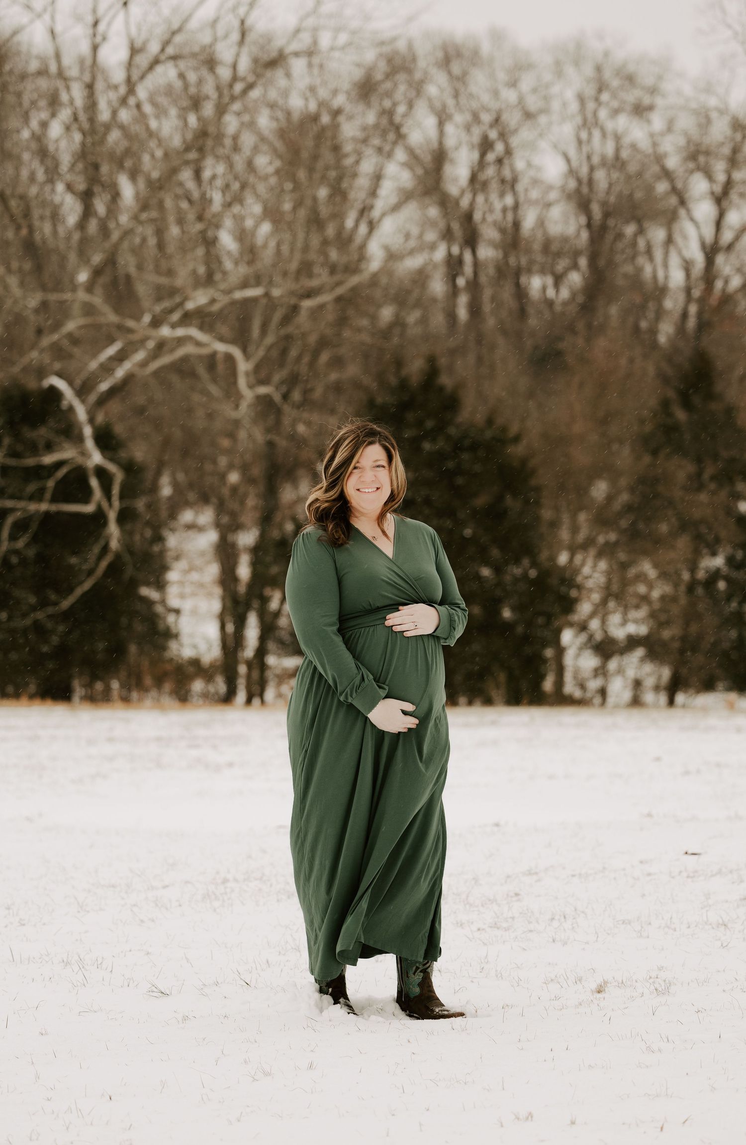A person in a long green sweater dress poses in a snowy winter landscape with bare trees in the background.