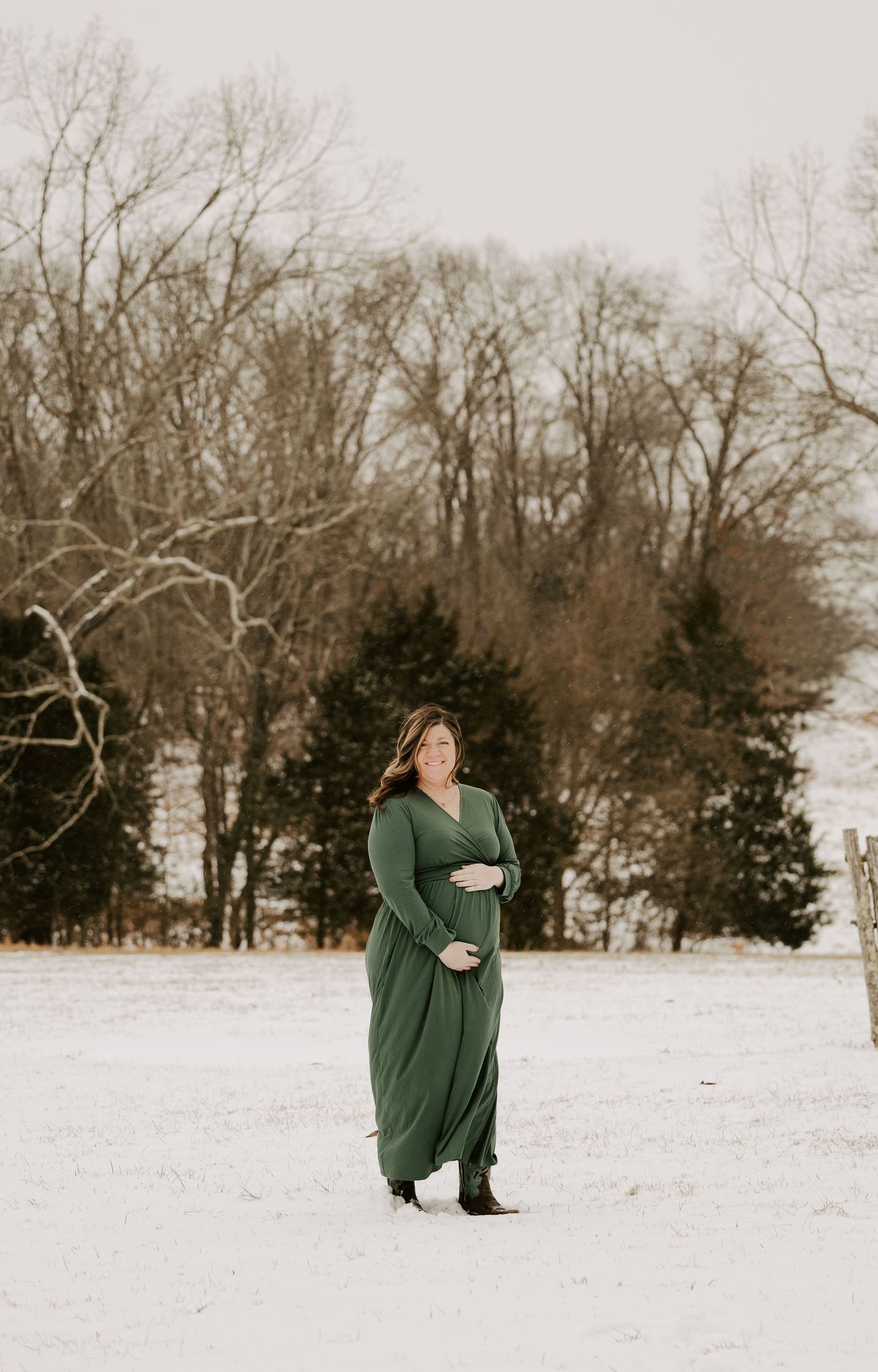 A figure in a dark green sweater stands in a snowy winter landscape with bare trees in the background.