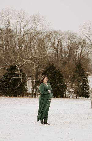 A person in a green coat walks through a snowy winter landscape with bare trees in the background.