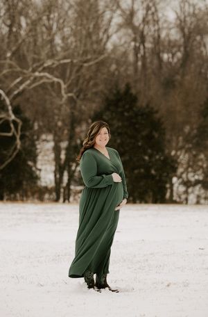 A person wearing a green sweater dress stands in a snowy winter landscape with bare trees in the background.