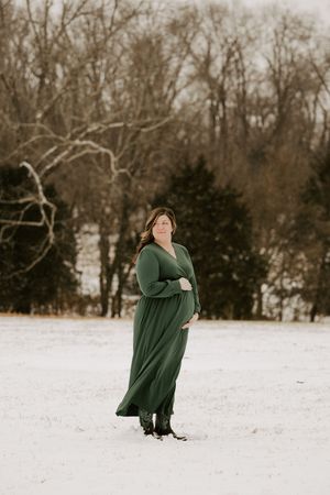 A person wearing a green sweater poses in a snow-covered field surrounded by winter trees.