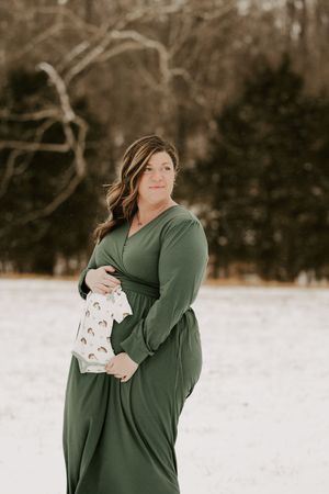 A pregnant woman in a green sweater poses in a snowy winter landscape holding a white sign.