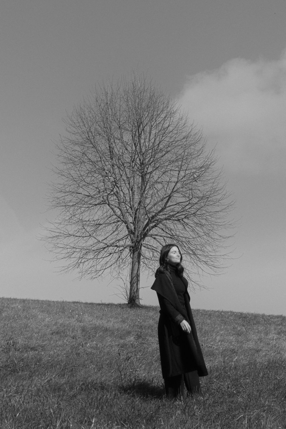 Black and white photo of a woman in a long coat looking up, standing near a bare tree on a grassy hill.