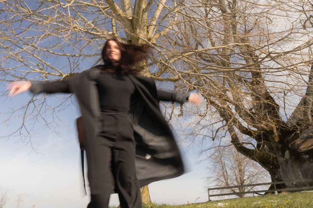 Woman in black coat spinning outdoors near bare trees on a bright winter day.
