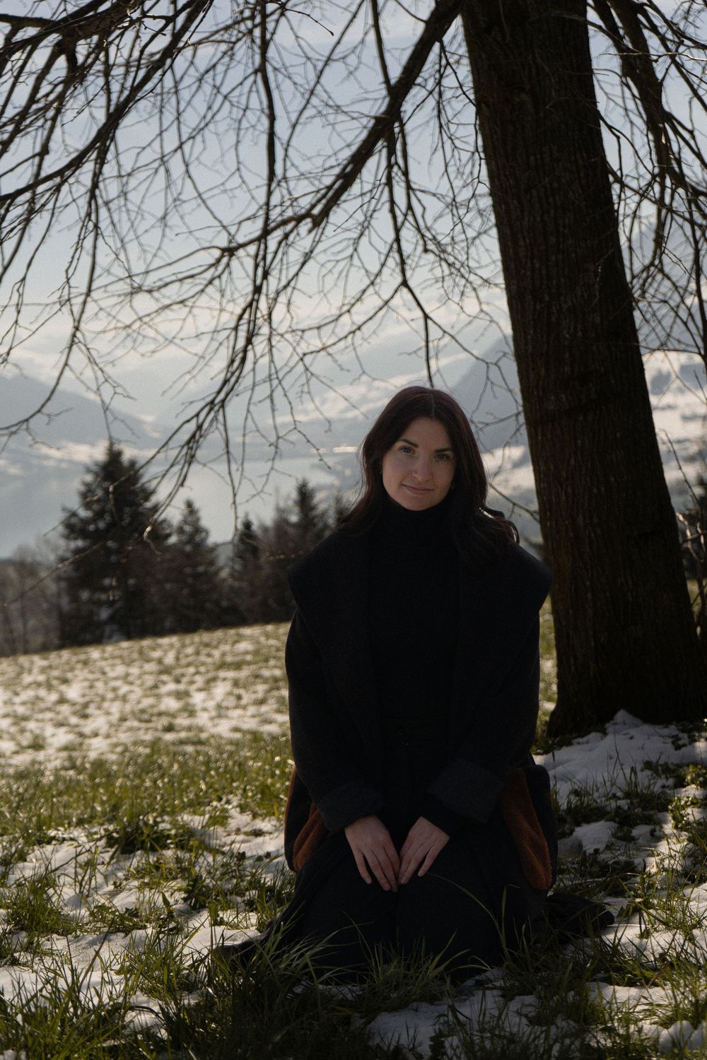Woman in black coat sitting by a bare tree on a snowy winter landscape with evergreens in the background.