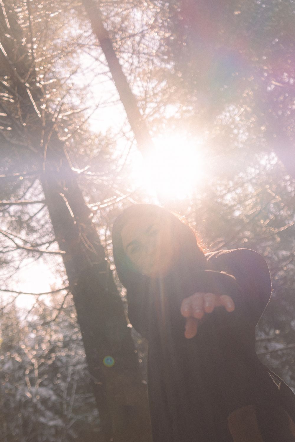 Person standing in a sunlit winter forest with lens flare and snow-covered trees in the background.