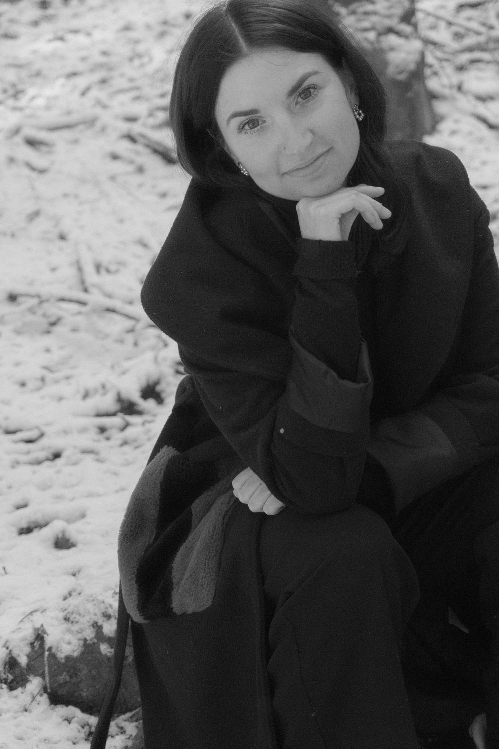 Black and white portrait of a dark-haired woman in a black coat sitting outdoors in snow, resting chin on hand.