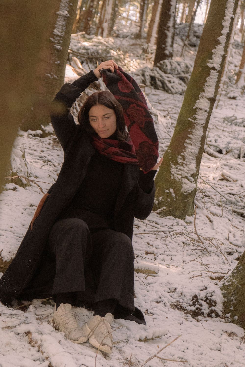 Woman in black coat holding red floral scarf in snowy winter forest, sitting among trees in white sneakers.