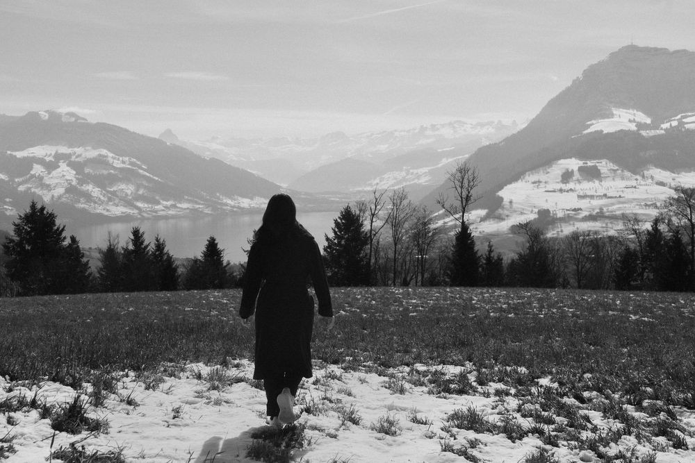 A lone figure in a dark coat walks through a snowy alpine meadow with mountain peaks in the background.