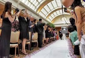 Wedding guests line both sides of an aisle covered in rose petals inside an elegant venue with arched glass ceiling.