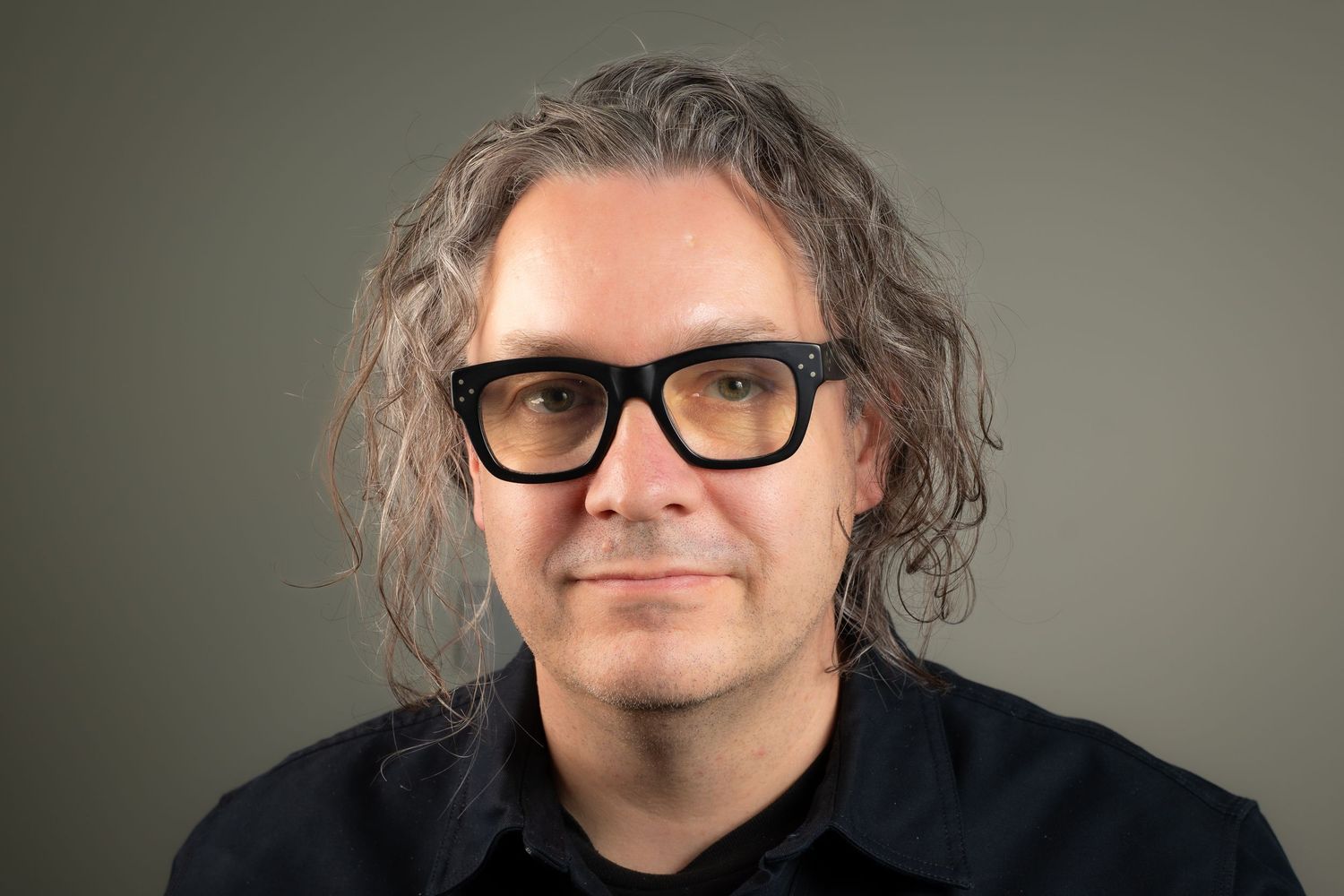 Keith with long brown hair and black thick-rimmed glasses wearing a dark shirt, professional headshot on gray background.