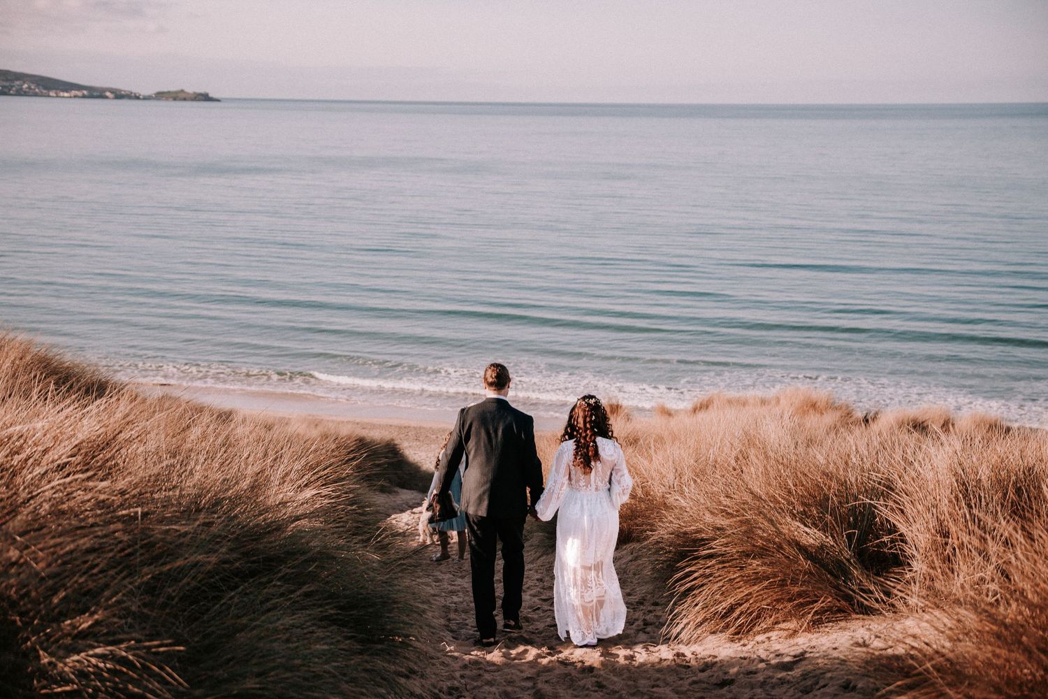 Wedding couple walks along scenic coastal beach at sunset with ocean waves and golden grass in view.