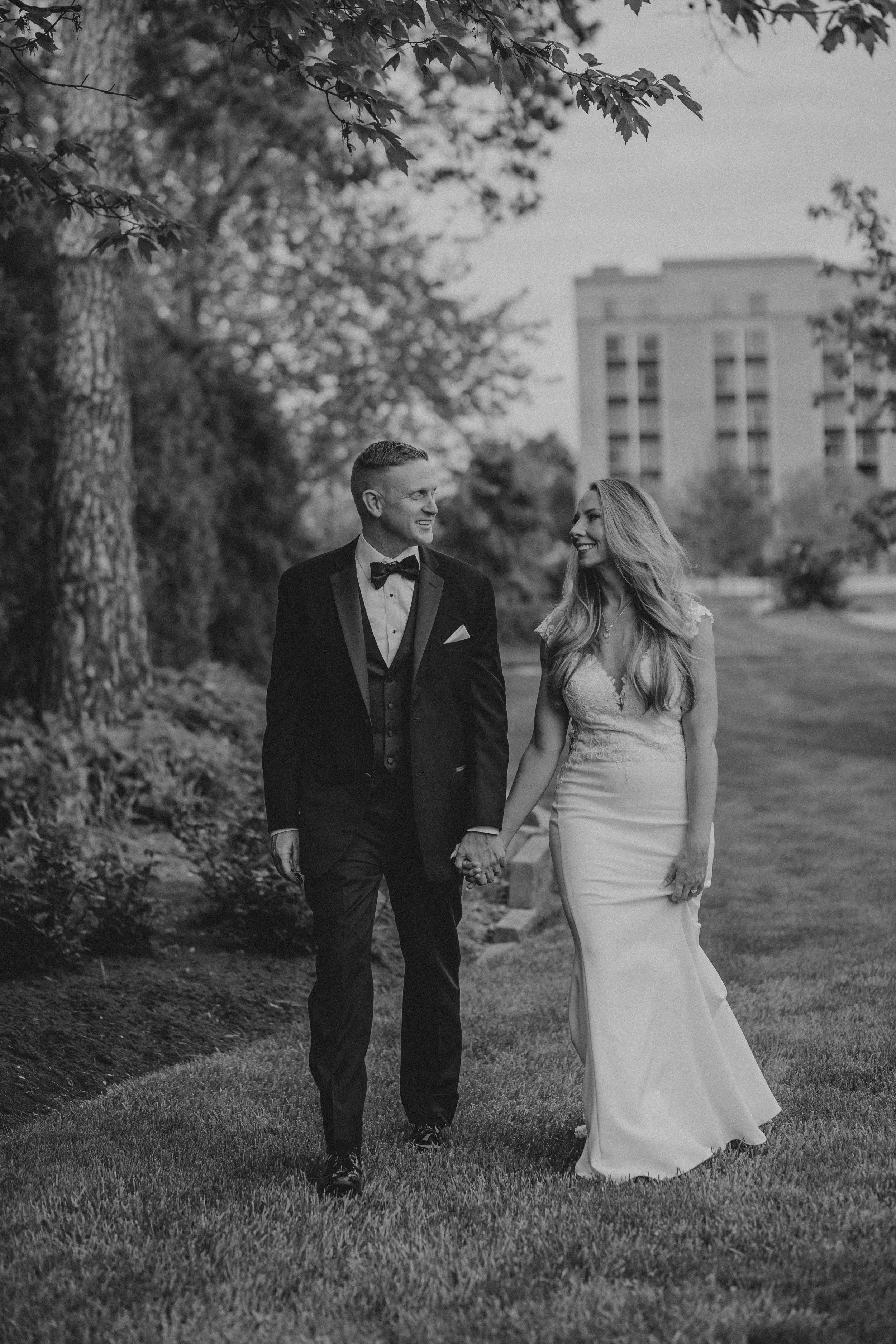 A romantic black and white wedding photo of a couple walking hand in hand outdoors with a building in the background.