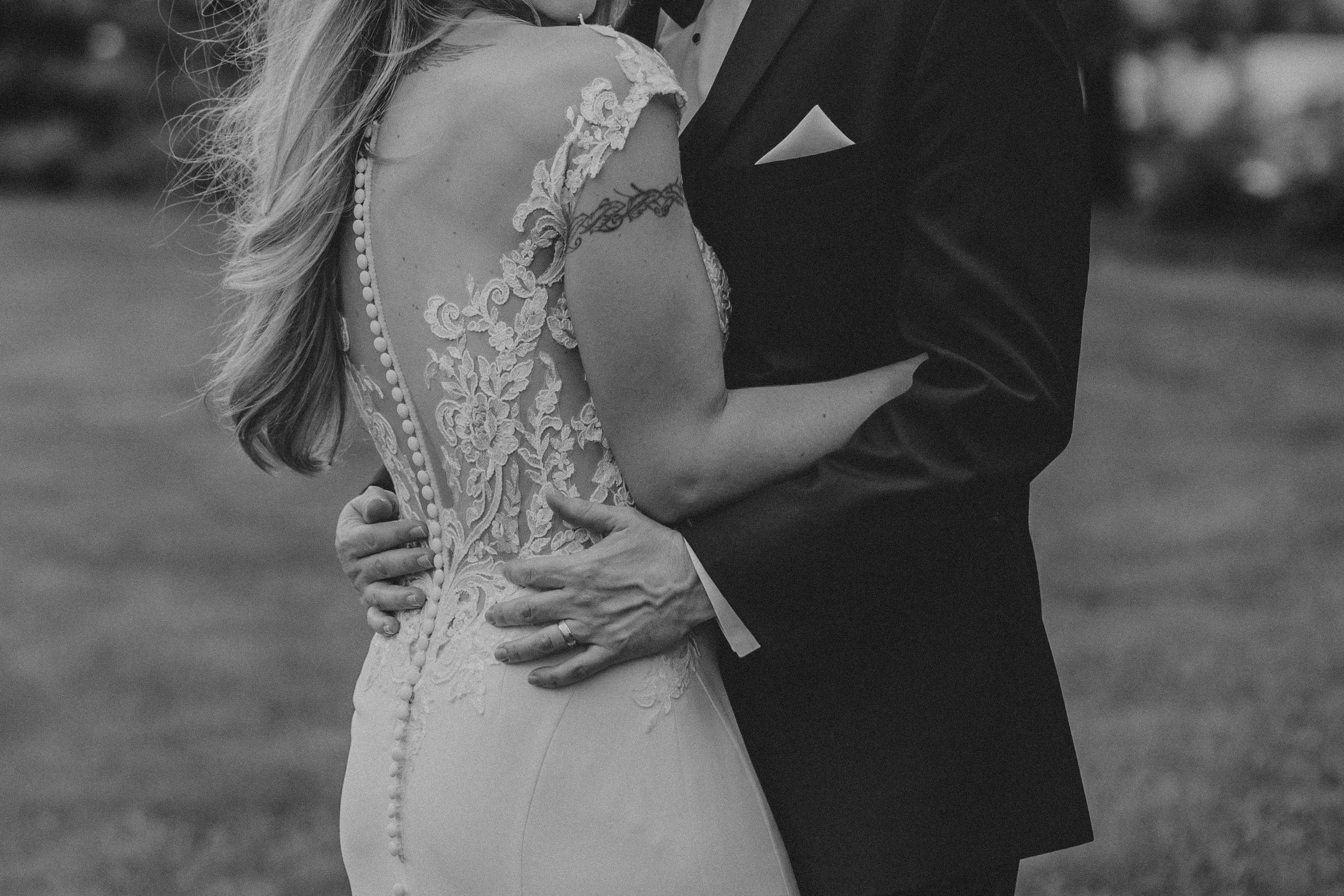 Black and white wedding photo showing intricate lace back detail of bridal gown as couple embraces outdoors.