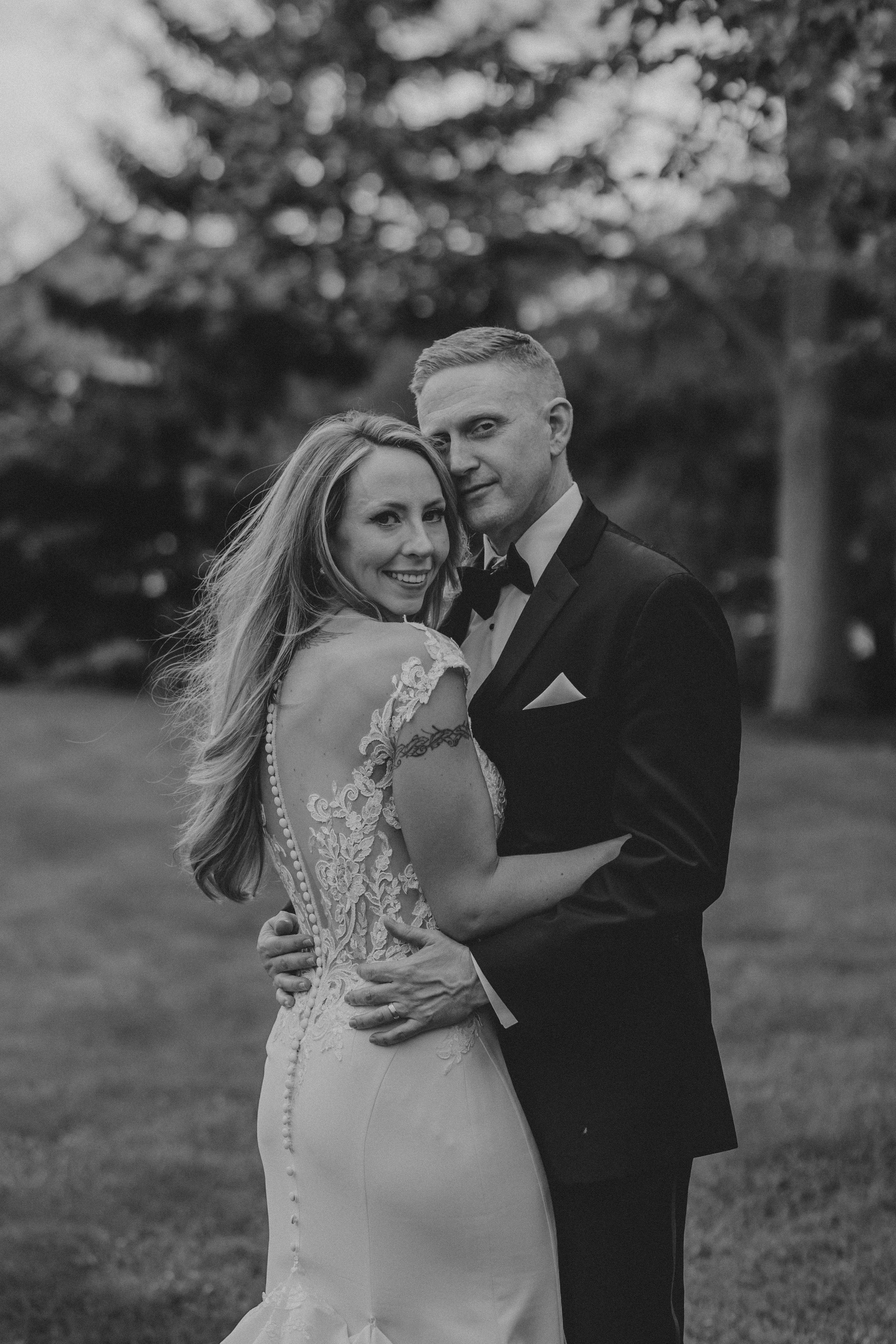 An intimate black and white portrait of newlyweds embracing outdoors, with the bride's detailed lace dress on display.