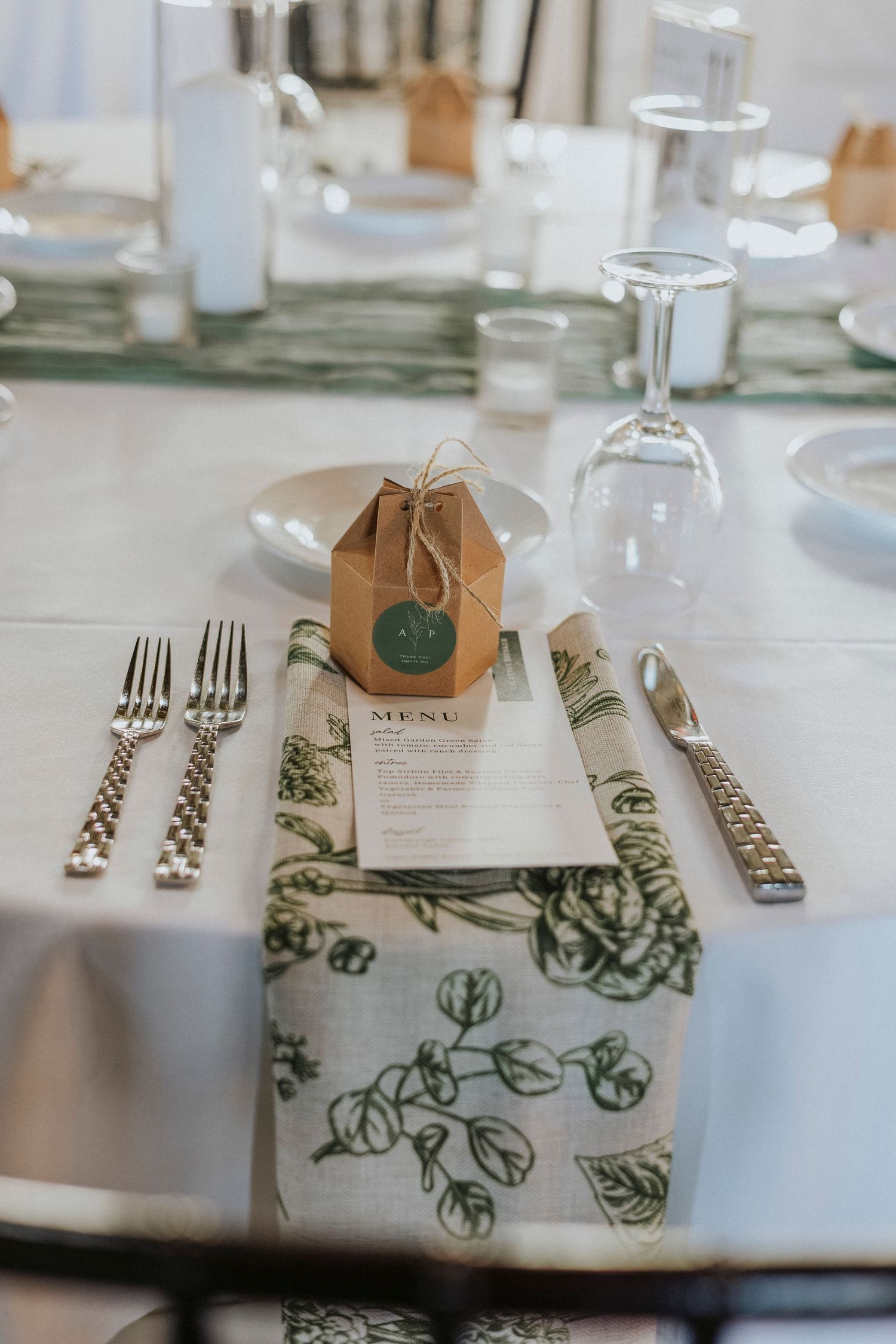 Wedding table settings with green floral placemats and brown favor boxes arranged elegantly on a white tablecloth.