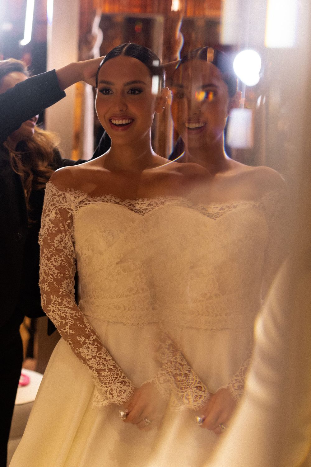 Two brides wearing elegant off-shoulder white lace wedding dresses stand together in a warmly lit indoor setting.