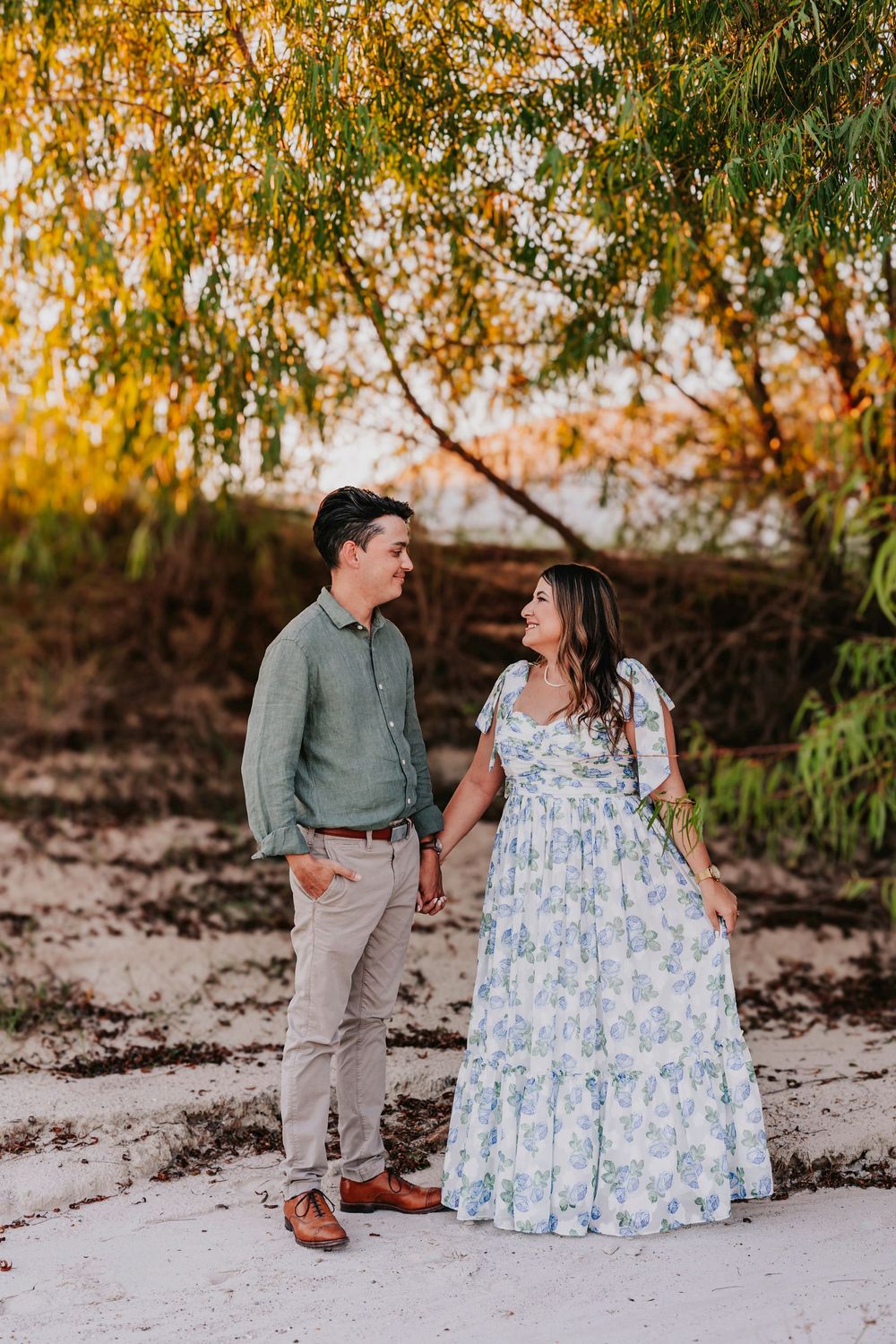 A couple in coordinated outfits stands together on white sand with autumn trees in background.