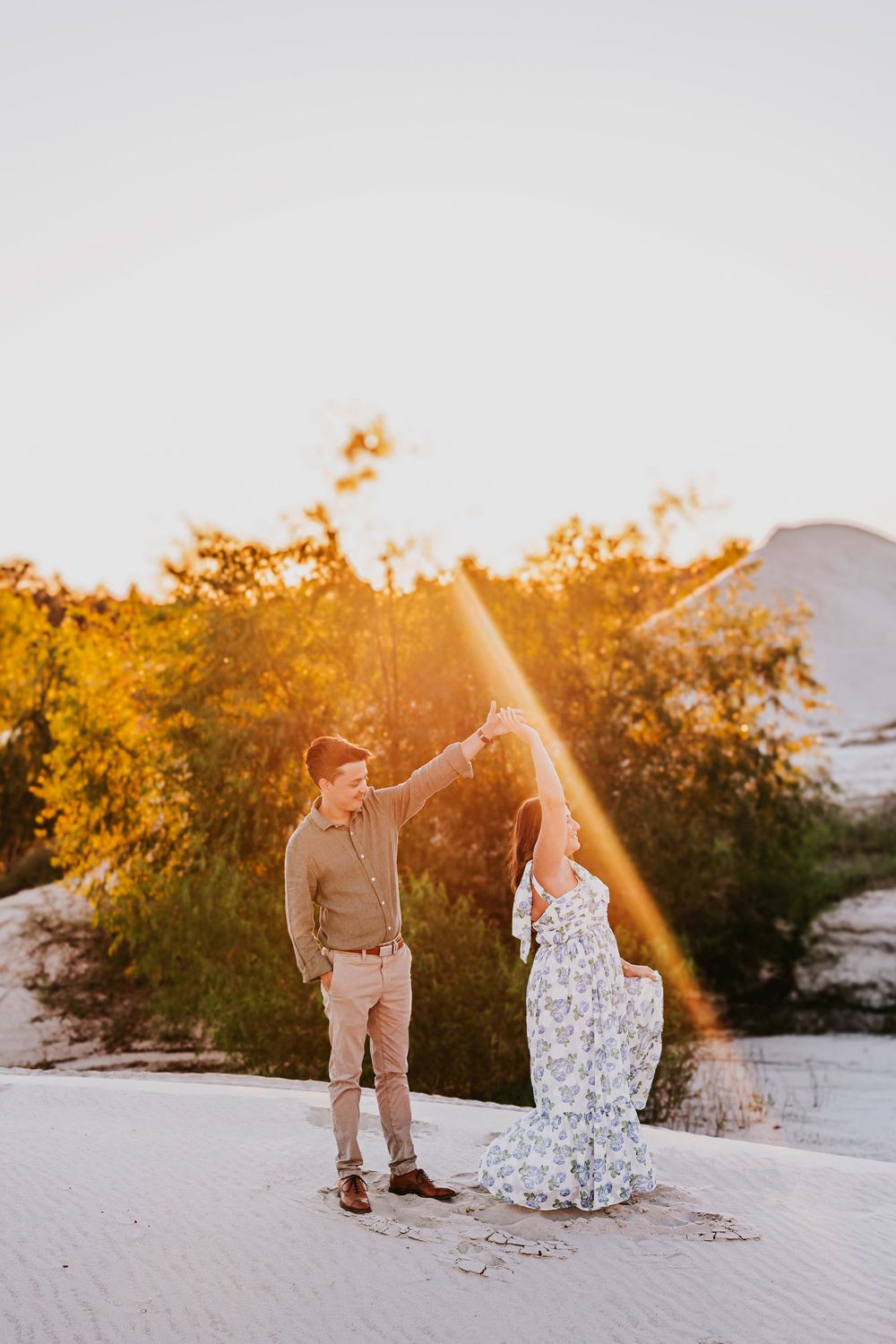 A couple poses romantically on white sand dunes at sunset with golden light filtering through trees.
