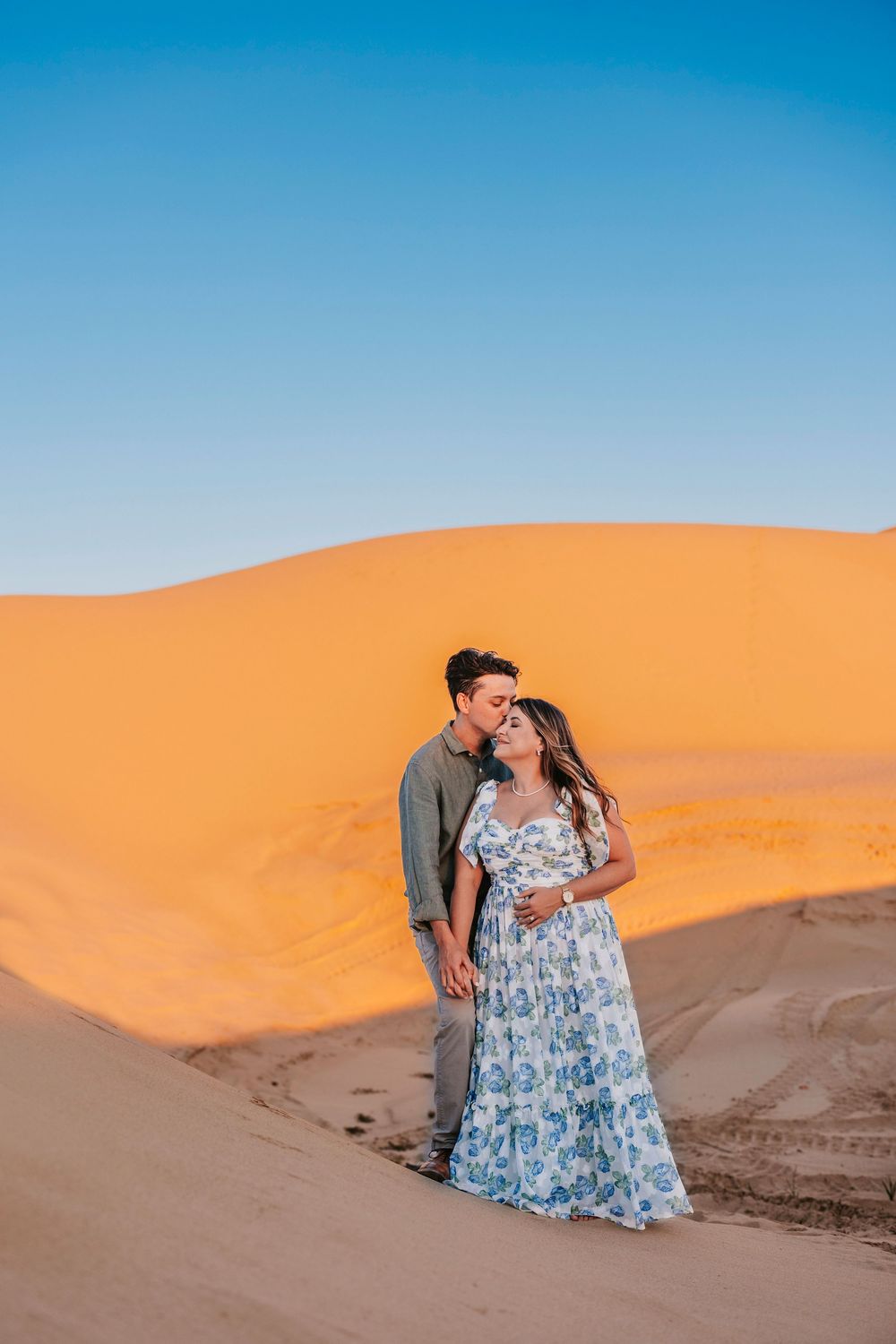 A couple stands together against vibrant orange sand dunes under bright blue sky.