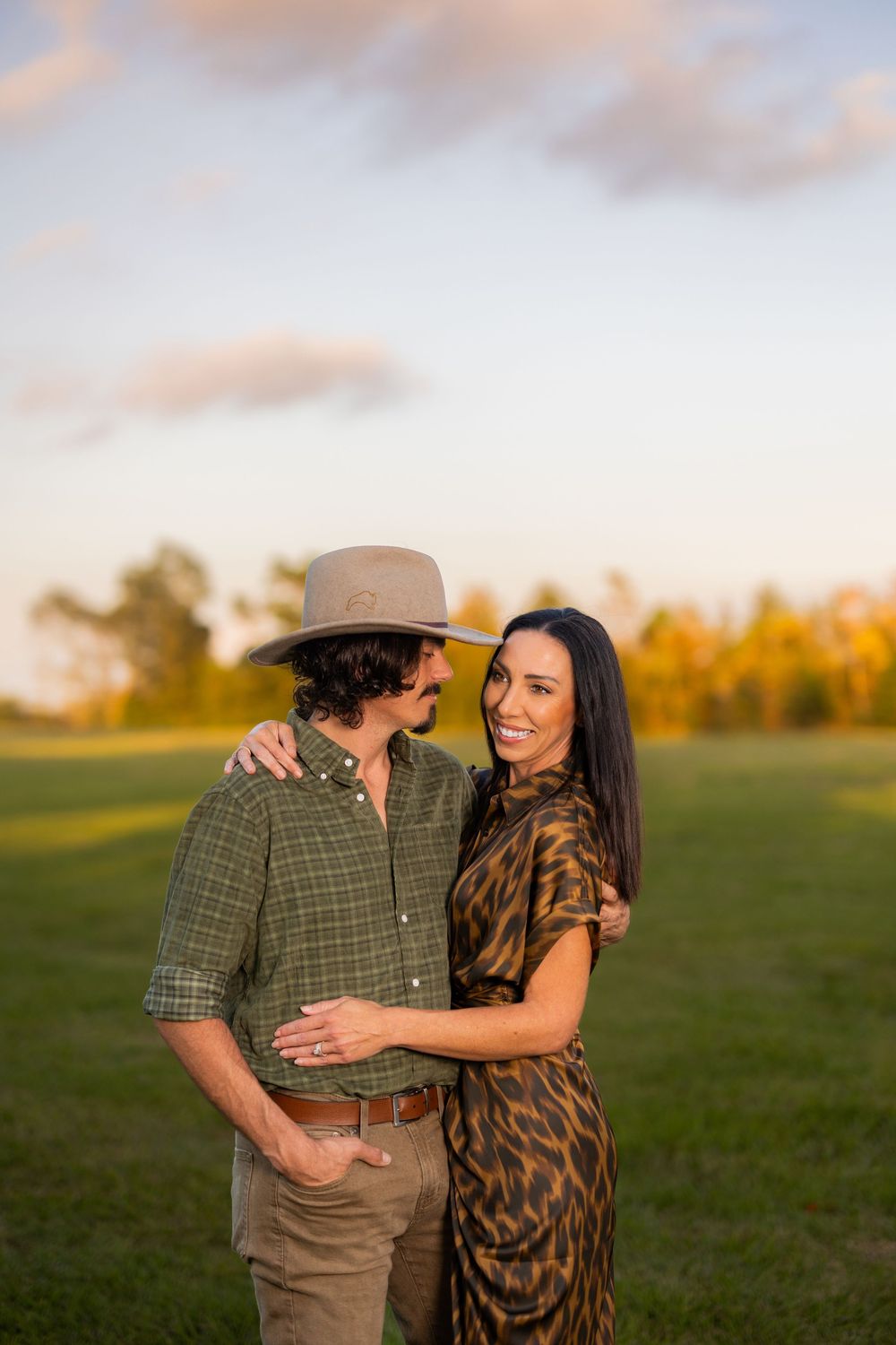A couple embraces in a golden field at sunset wearing a plaid shirt and animal print dress.