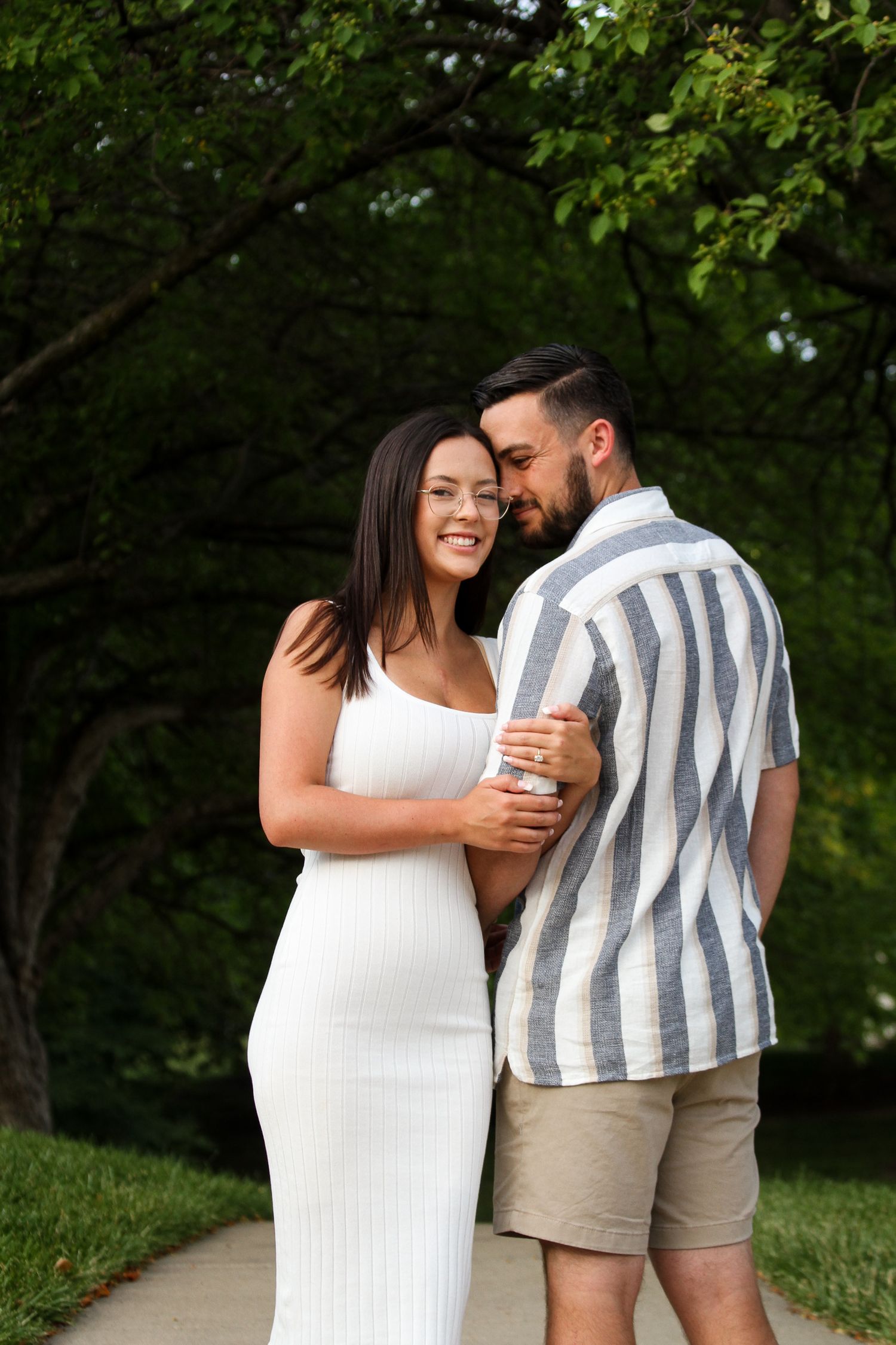 A woman smiles softly for the camera as her fiance leans into her under a dramatic canopy of leafy green trees during an engagement session.