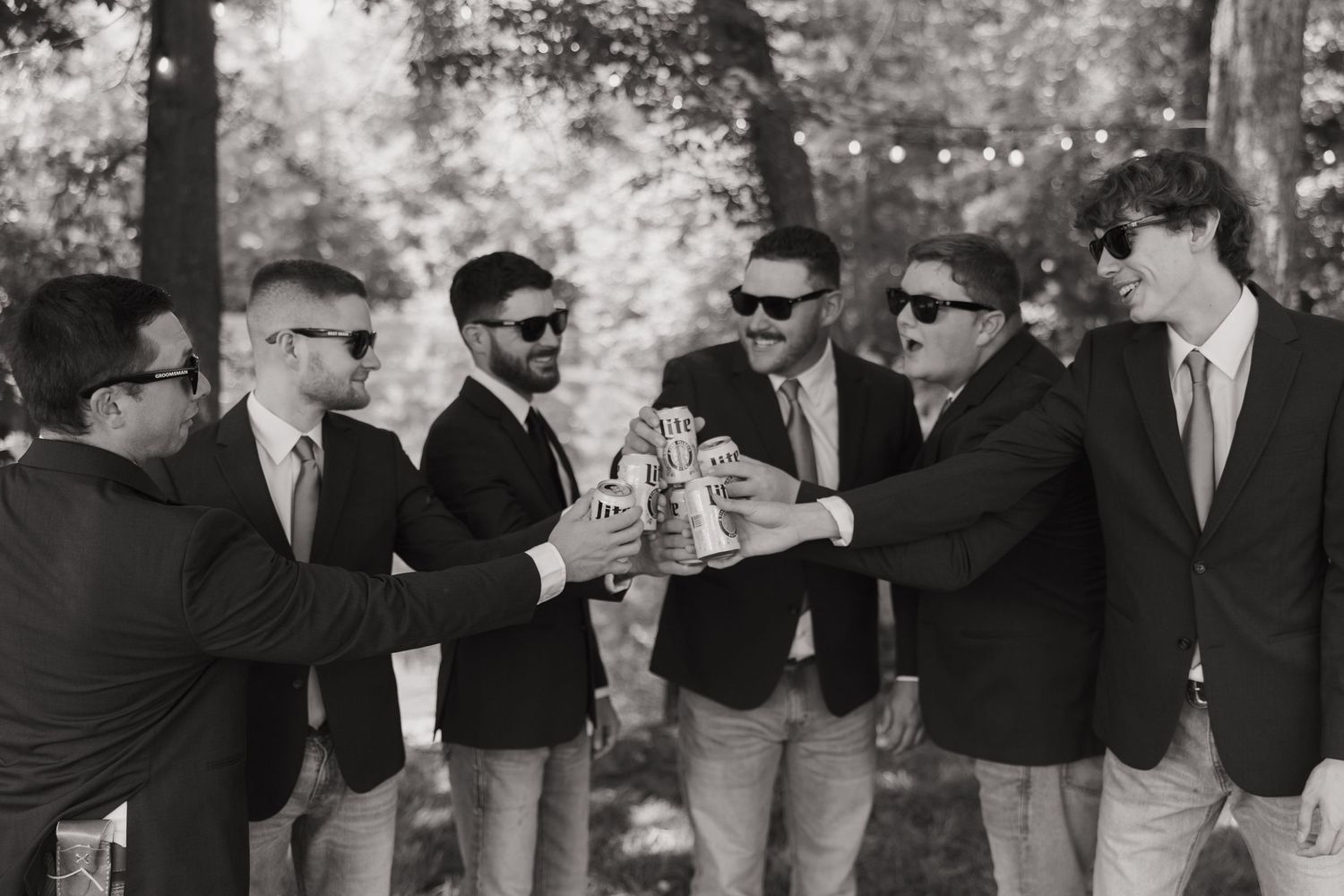 A black and white photo shows a line of people dancing with their hands on each others shoulders at an outdoor event.