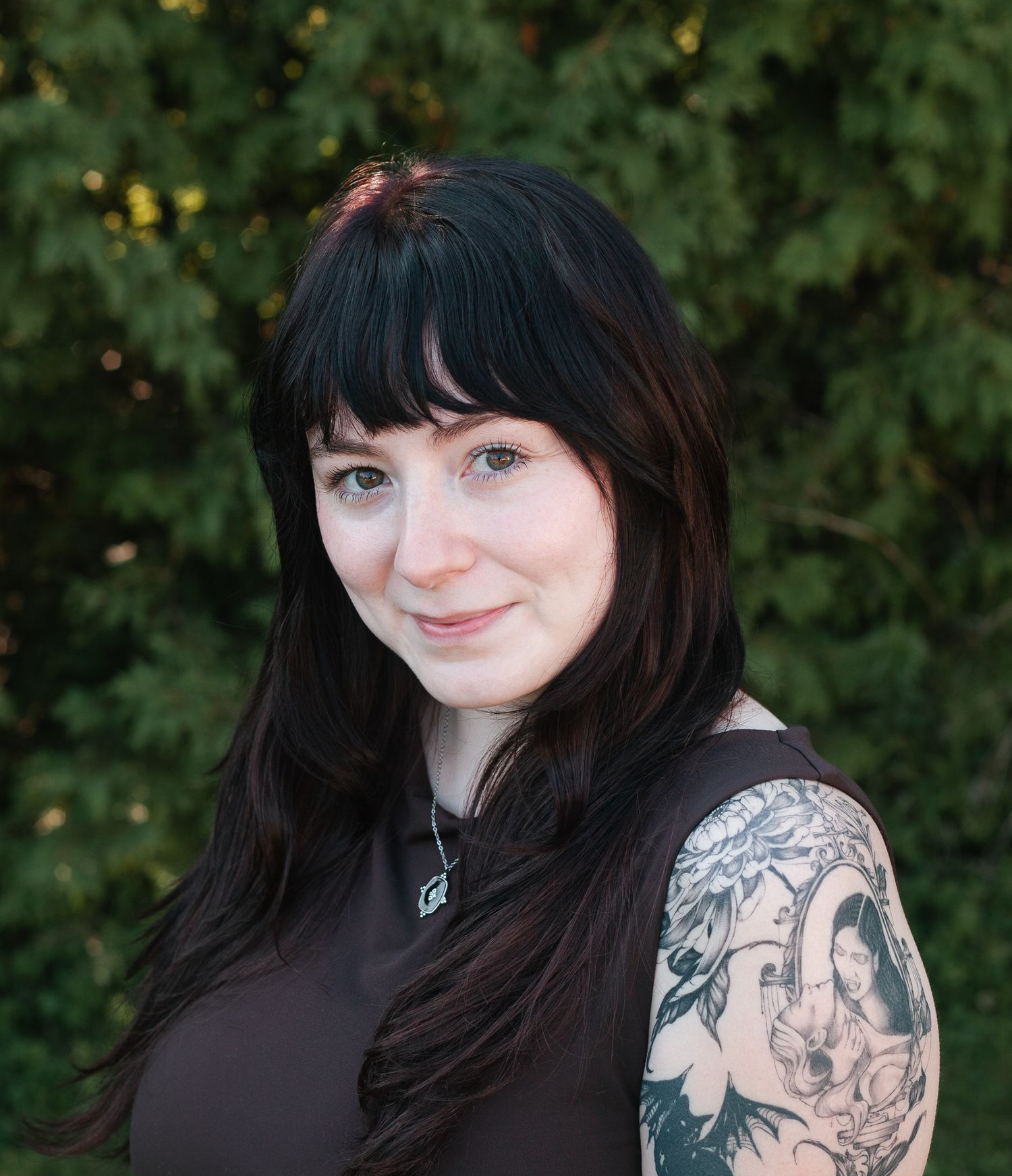 Young woman with long black hair and floral arm tattoo smiling outdoors near green foliage.