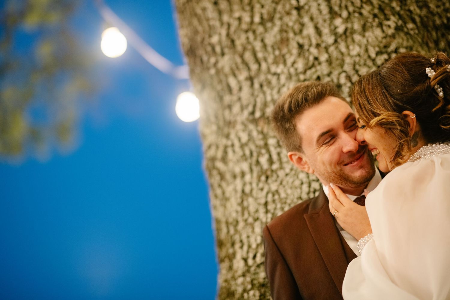 A romantic evening moment captured under string lights and a large tree trunk with a soft blue sky backdrop.