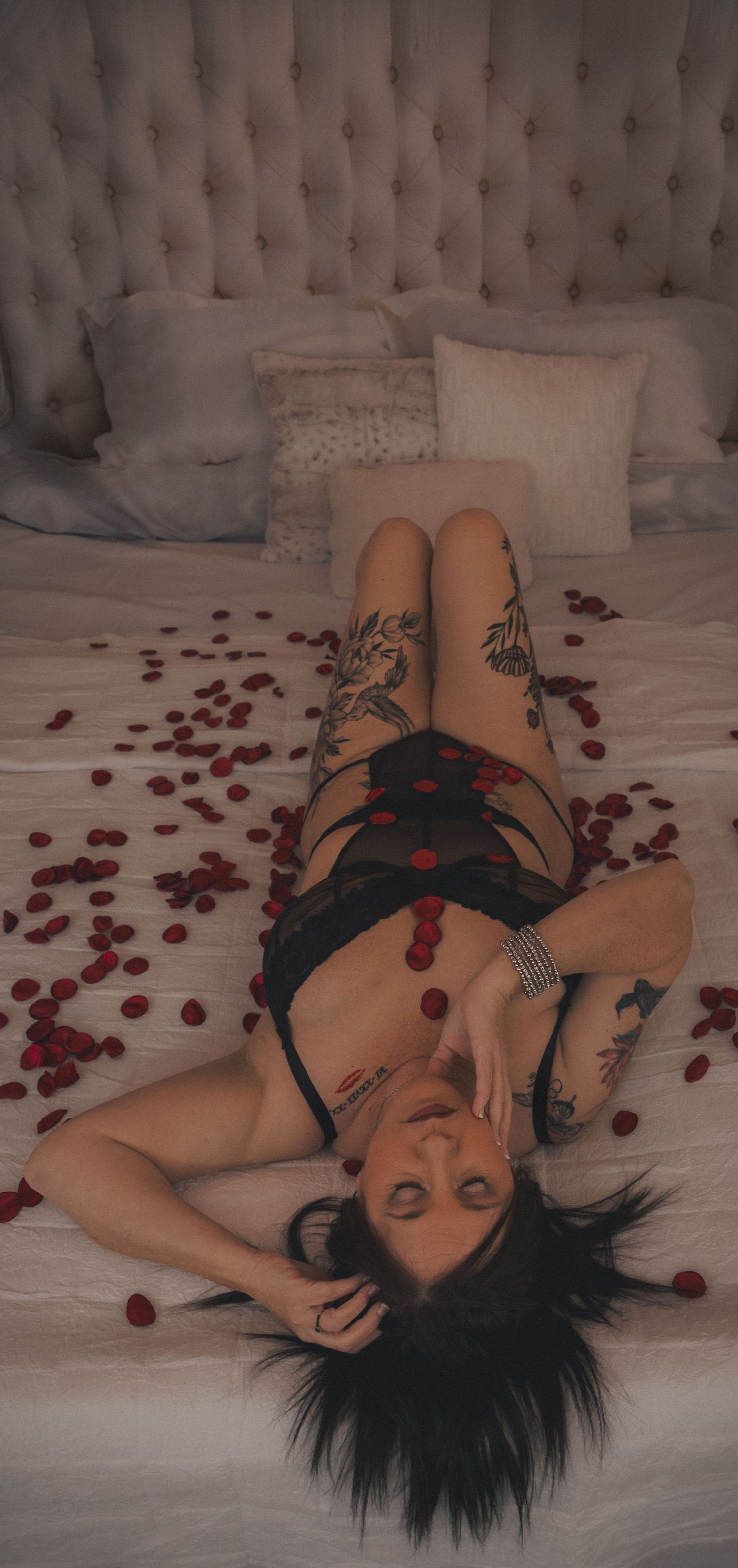 Woman in black lingerie lying on white bedding with scattered red rose petals in artistic boudoir photography.