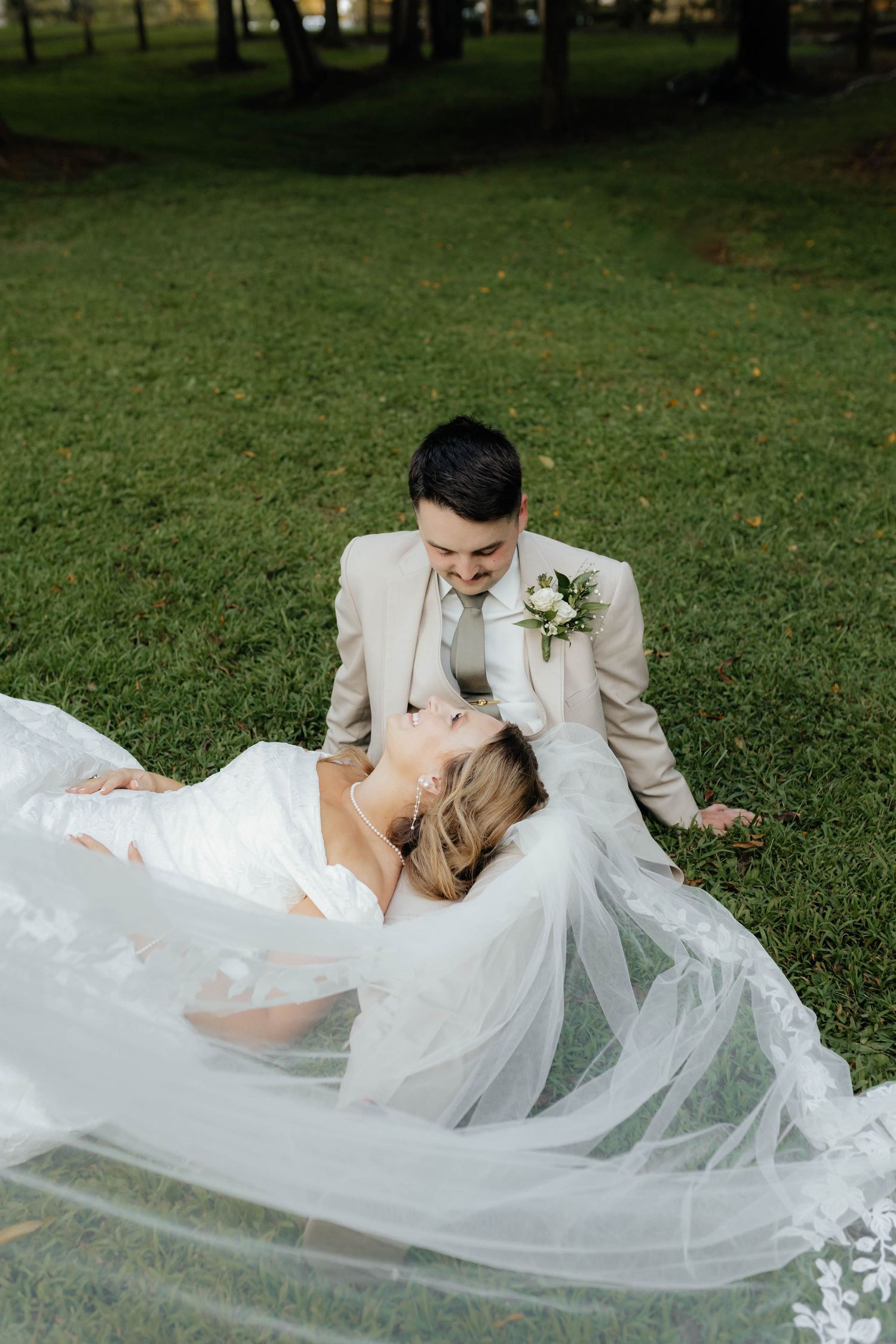 A romantic wedding couple lies on green grass as the bride's white veil flows gracefully in the breeze.