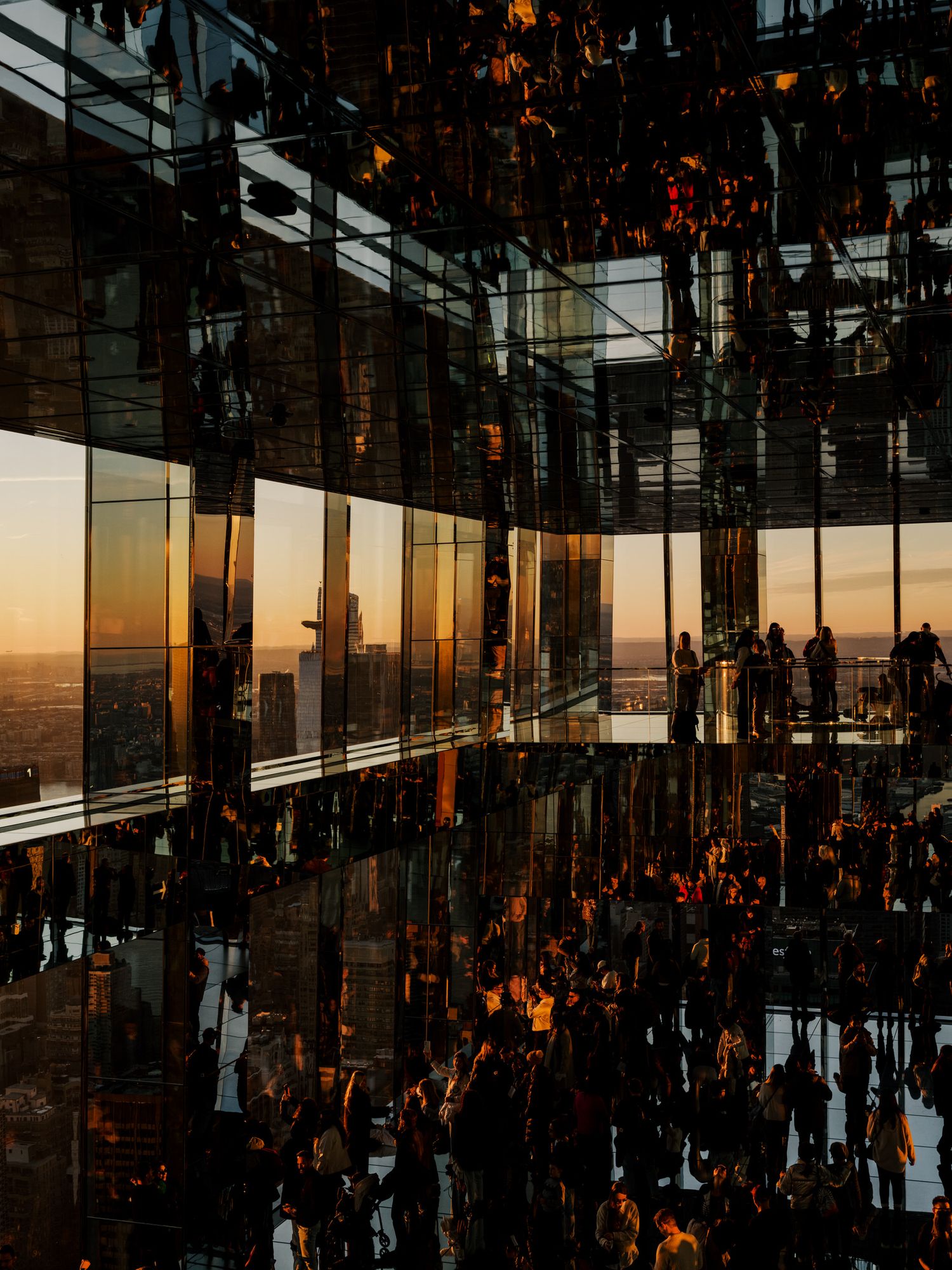 Silhouetted visitors enjoy sunset views through floor-to-ceiling windows at an observation deck in a modern skyscraper.