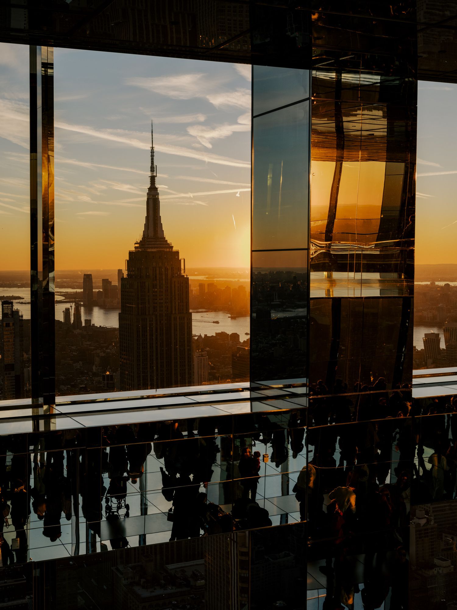Sunset silhouettes the Empire State Building in New York City with reflective glass windows and people viewing the skyline.