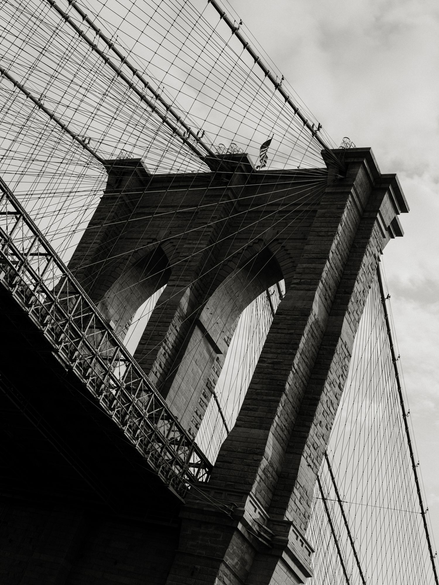 Dramatic black and white low angle view of Brooklyn Bridge's architectural details and cables against cloudy sky.