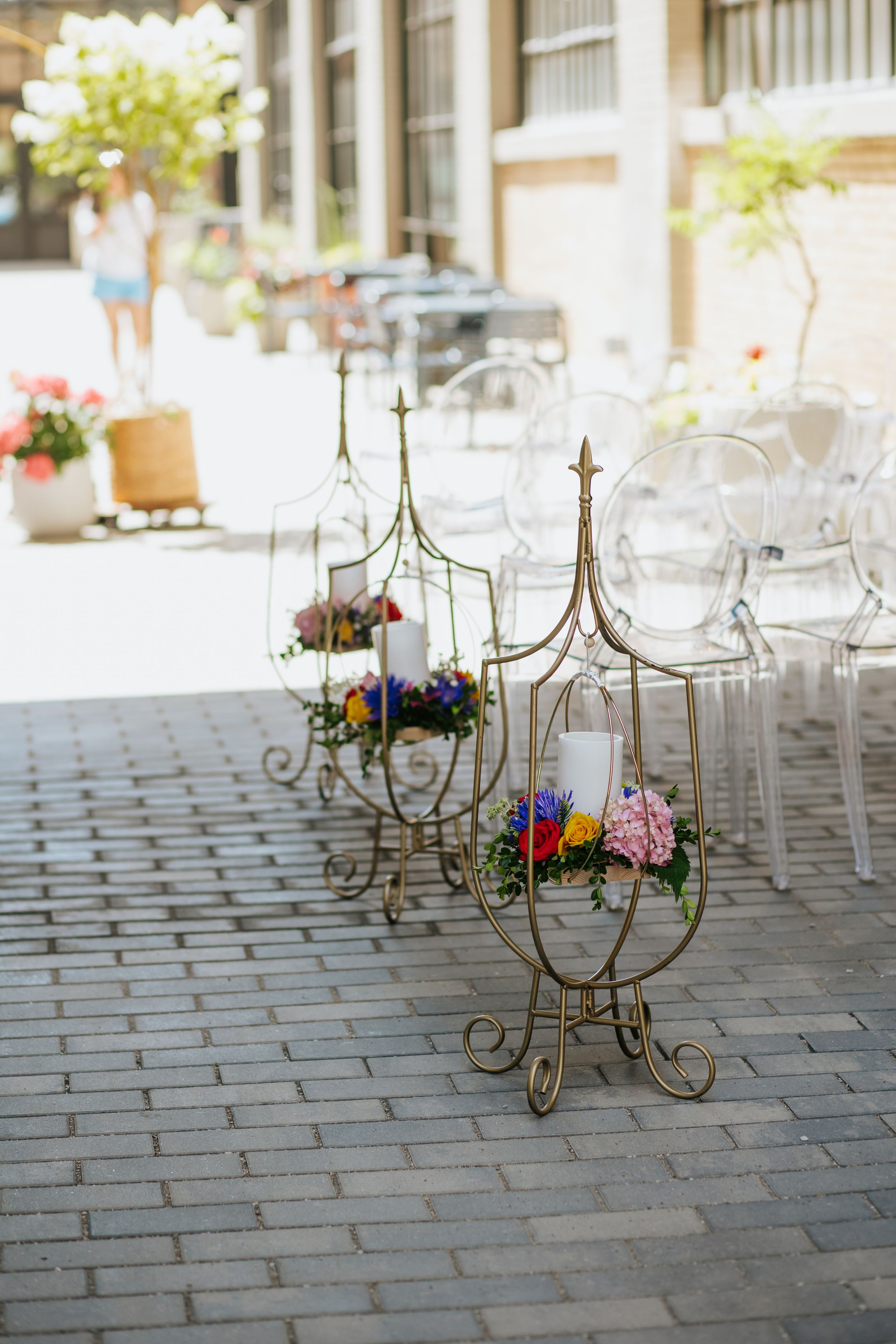 Decorative gold candelabra stands arranged on a brick patio with white chairs and colorful floral arrangements.