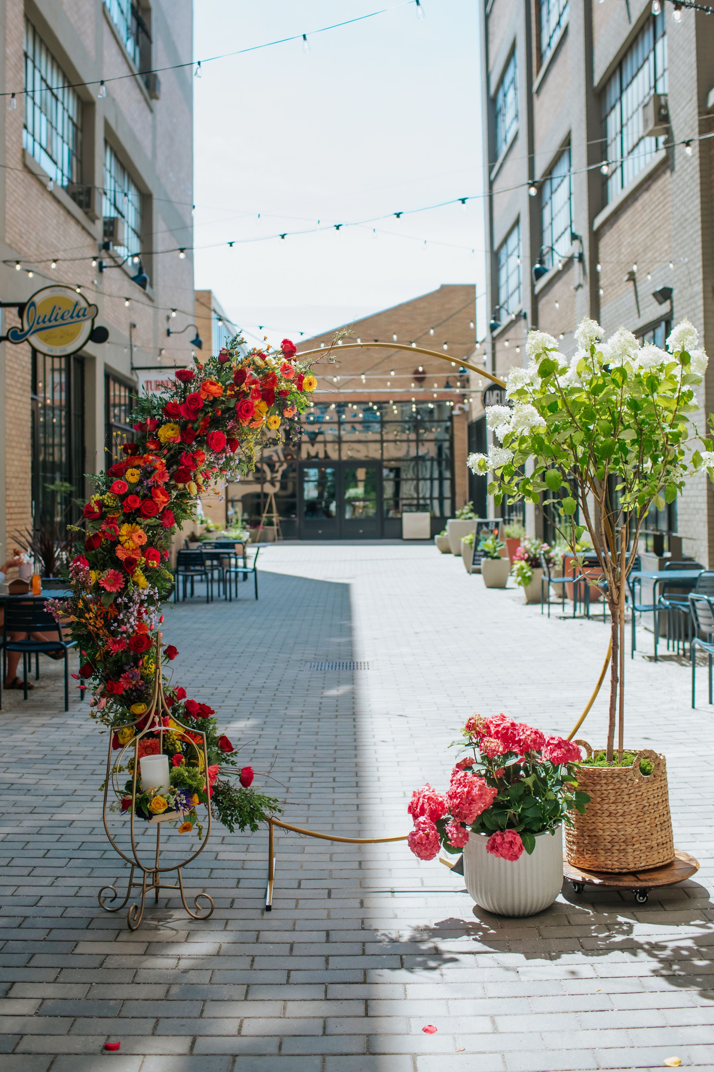 A series of floral arrangements with red and orange blooms in decorative planters line a brick paved walkway.