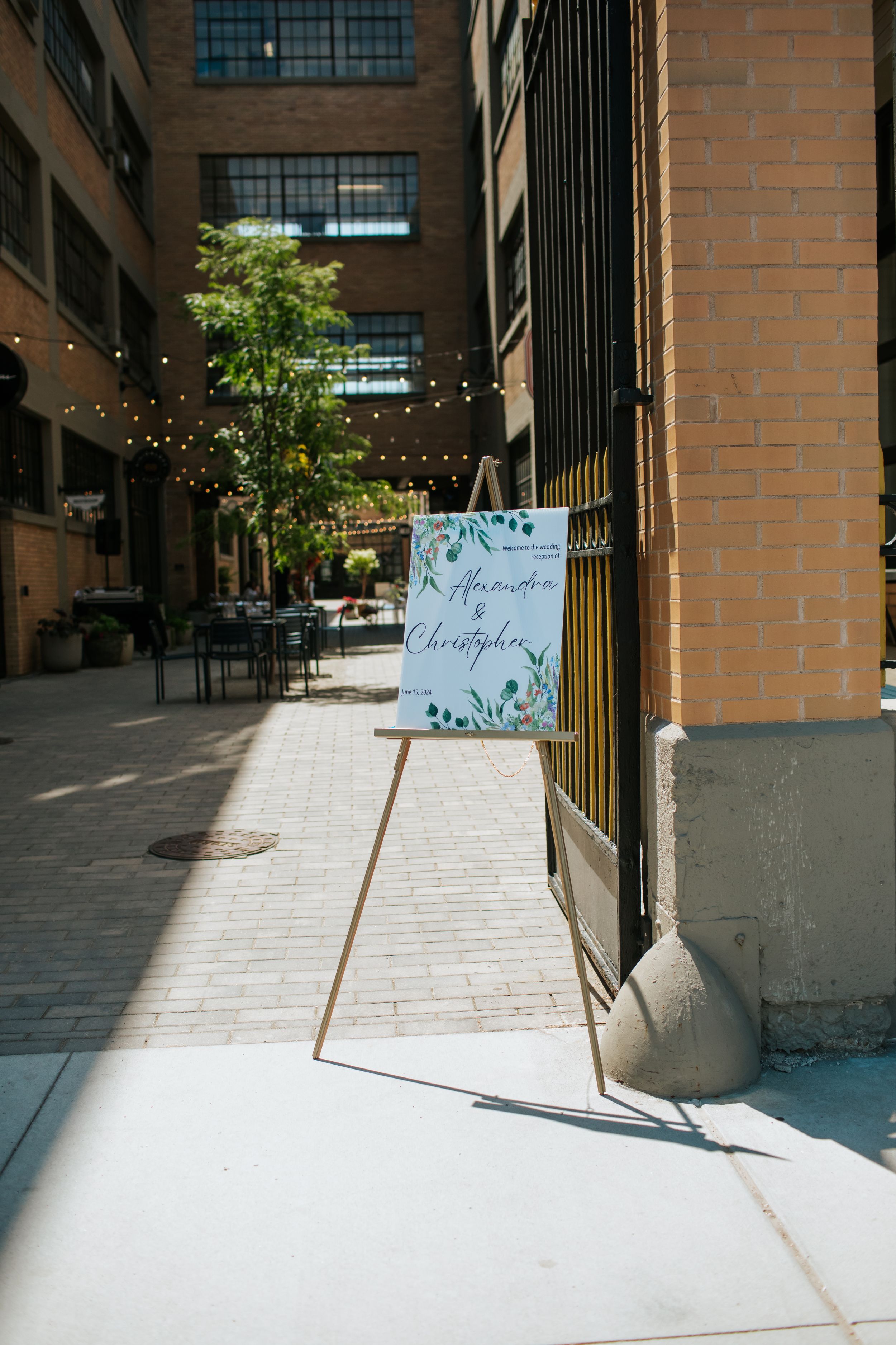 A wedding welcome sign stands in a sunlit brick hallway with string lights and greenery decorations.