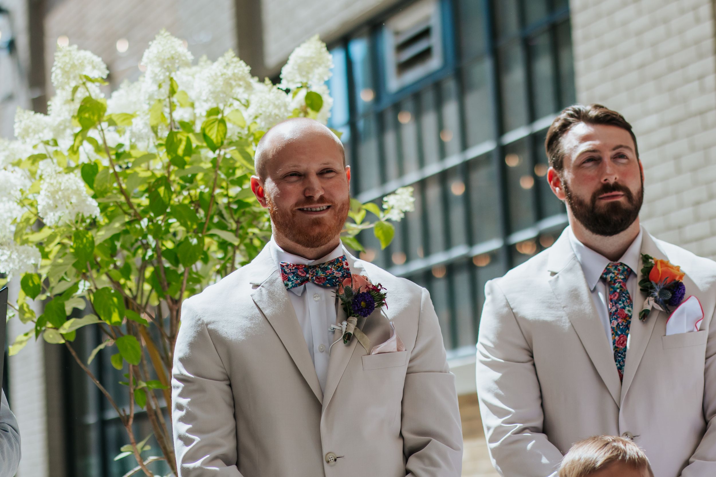 Two groomsmen in light beige suits with floral ties stand at an outdoor wedding ceremony with white flowers nearby.