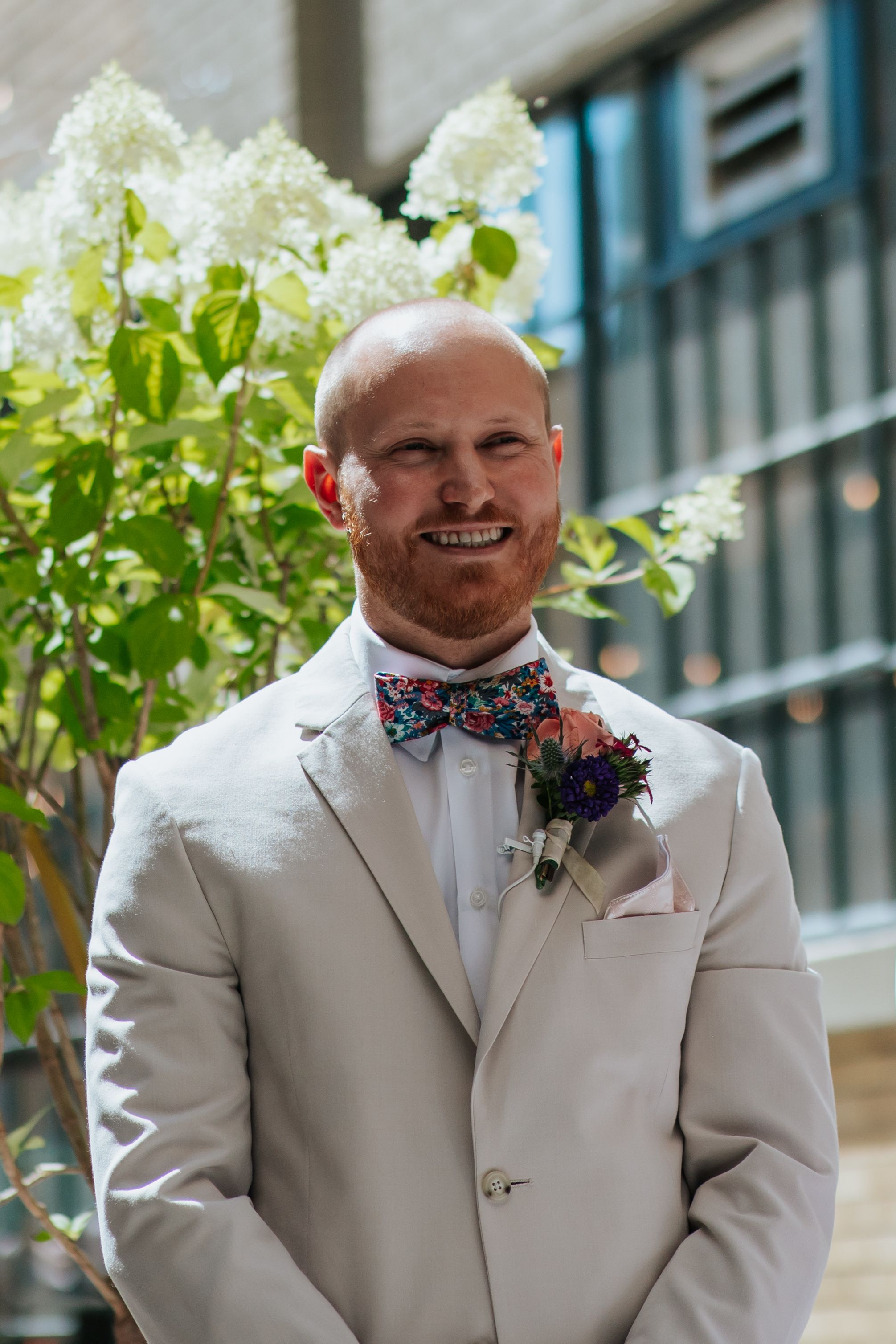 A person in a light beige suit and floral bow tie smiles outdoors with greenery in the background.